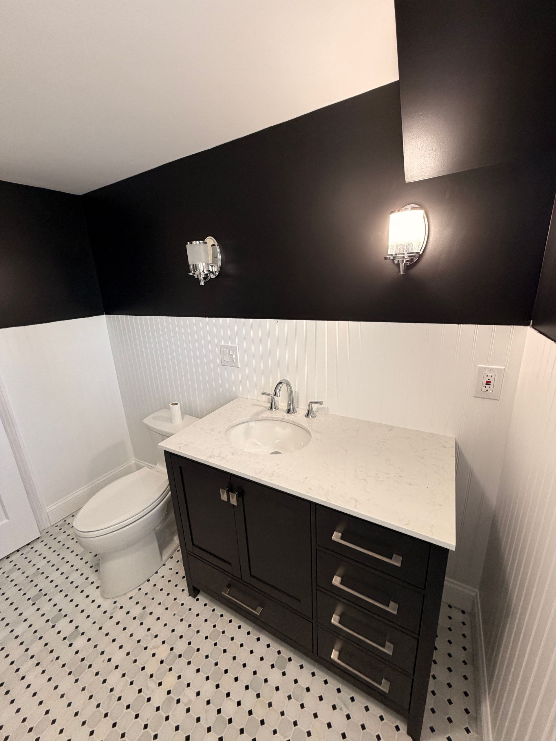 Black-and-white bathroom with pedestal sink, dark vanity, toilet, and patterned tile floor.