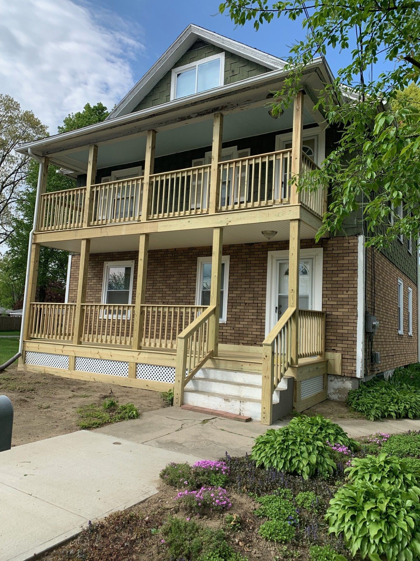 Two-story house with wood balconies and steps; brown siding, green trim, and garden in front.