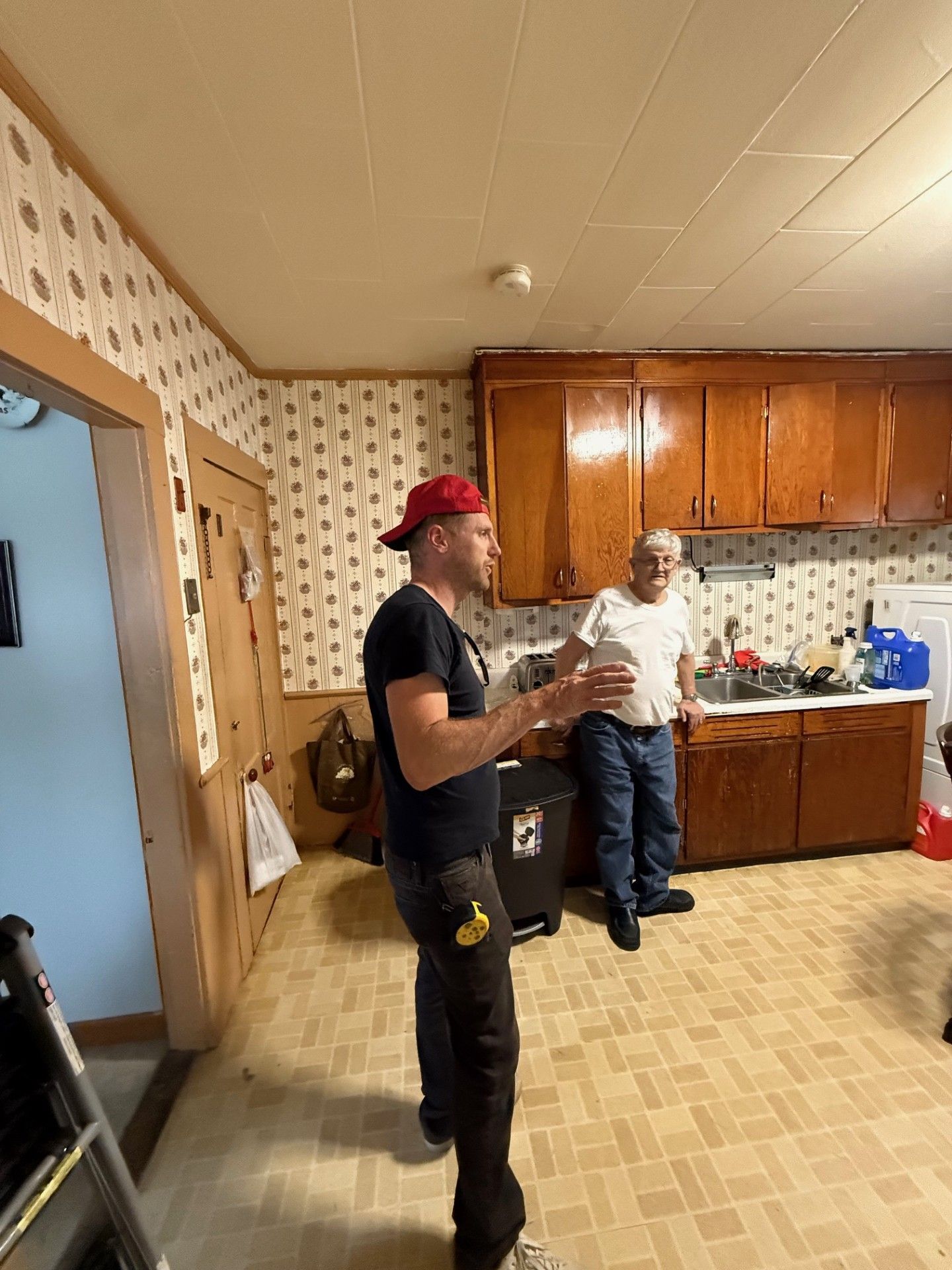 Man in red cap and another man in a kitchen, discussing something near cabinets, a door, and a ladder.