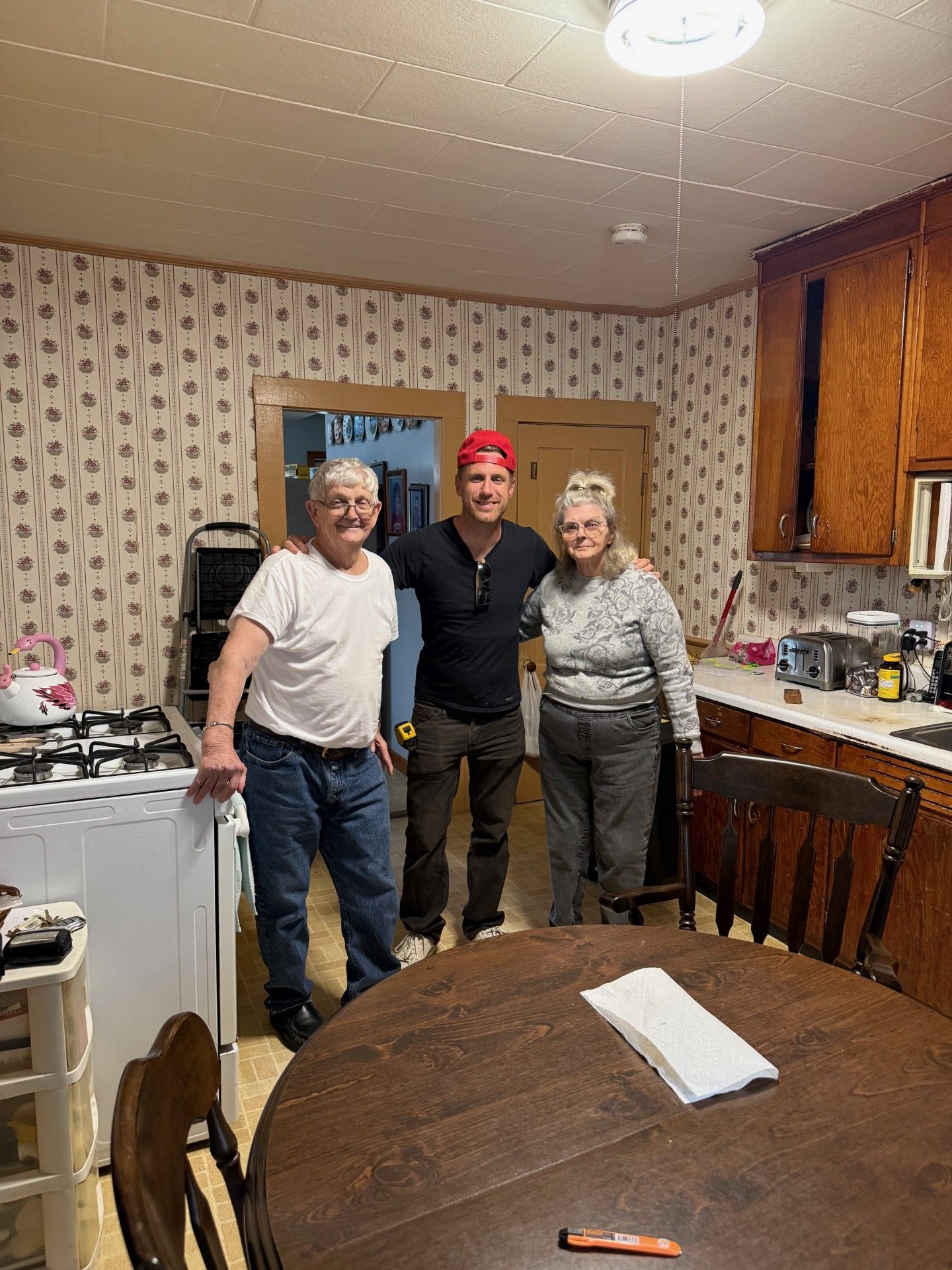 Three people pose in a kitchen: an older man, a younger man with a red bandana, and an older woman.