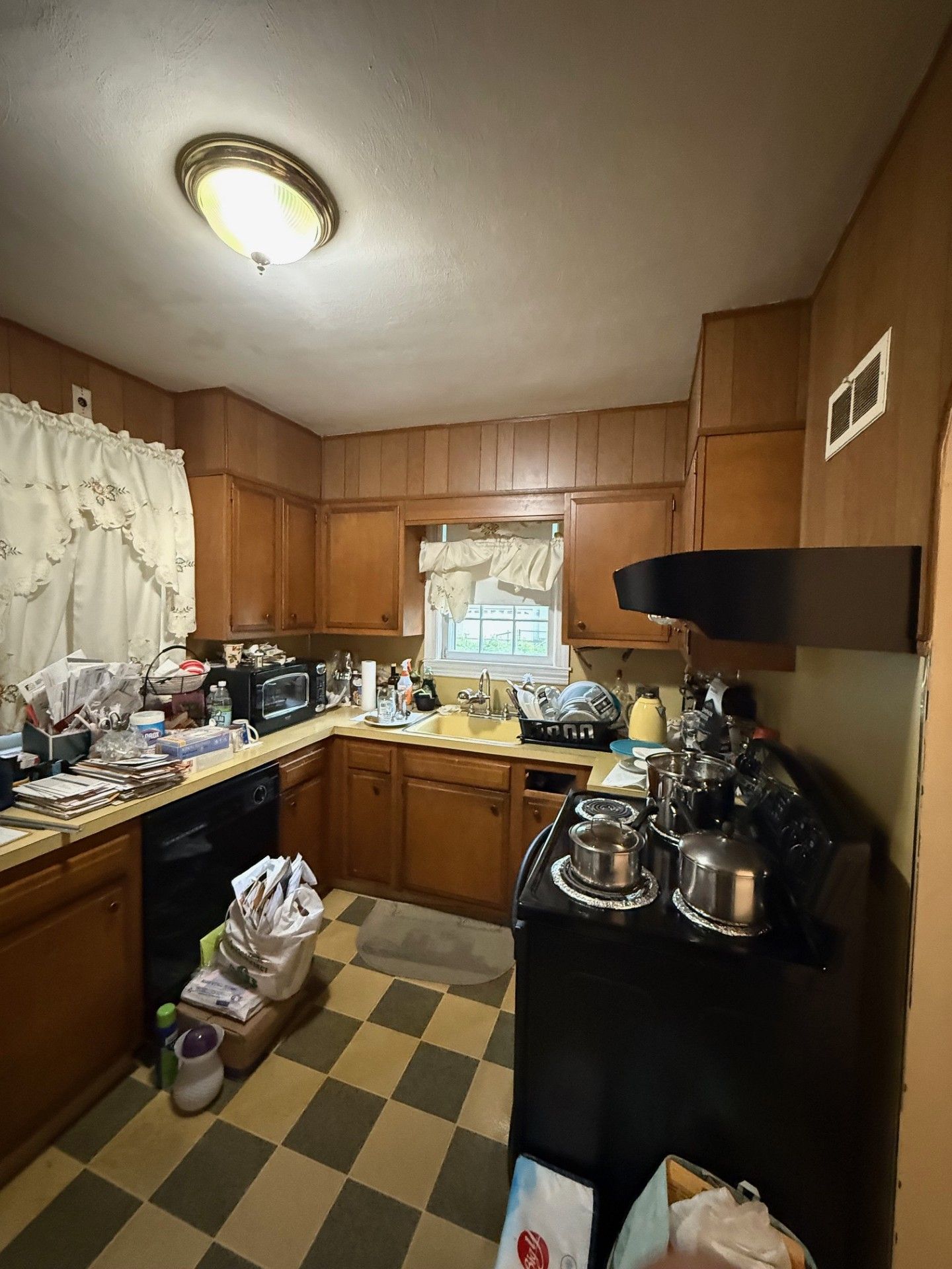 A cluttered kitchen with wooden cabinets and appliances, including a black stove and a window with a curtain.