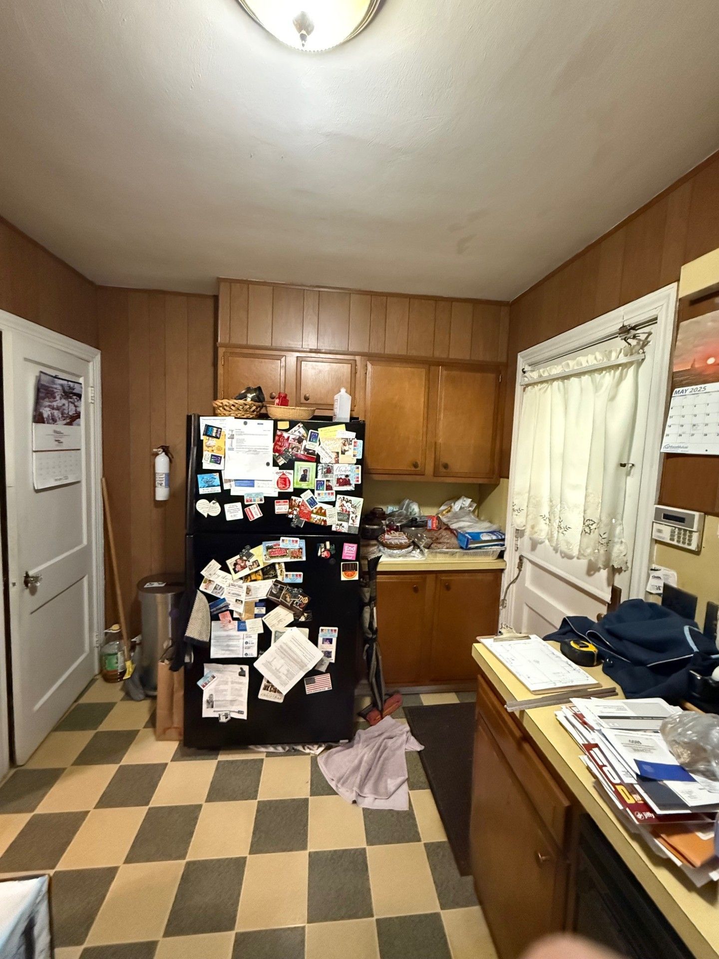 A cluttered kitchen with wood cabinets, refrigerator covered in notes, and checkered floor.