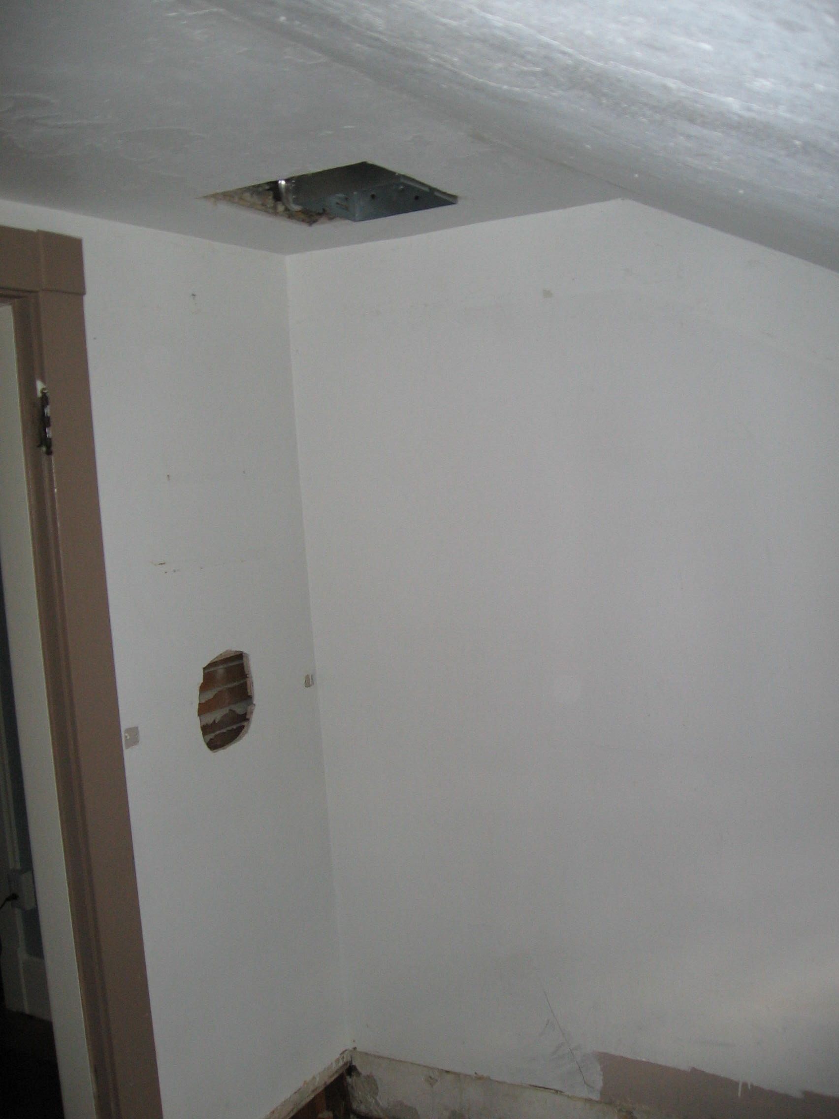 Corner of room with damaged wall, ceiling vent, and doorway. Walls are white.