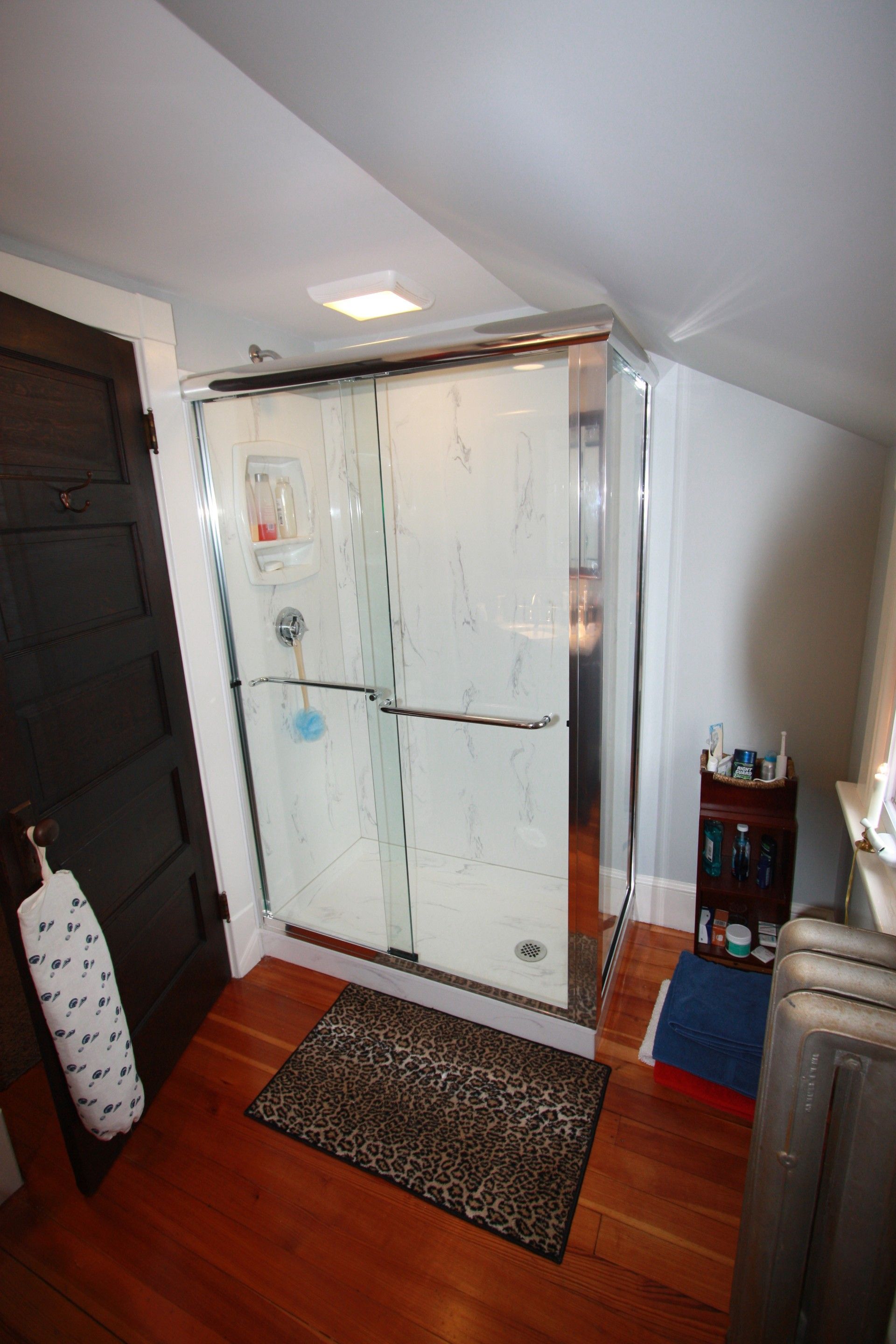 Bathroom with angled ceiling, glass shower, dark door, hardwood floor, and a small leopard rug.