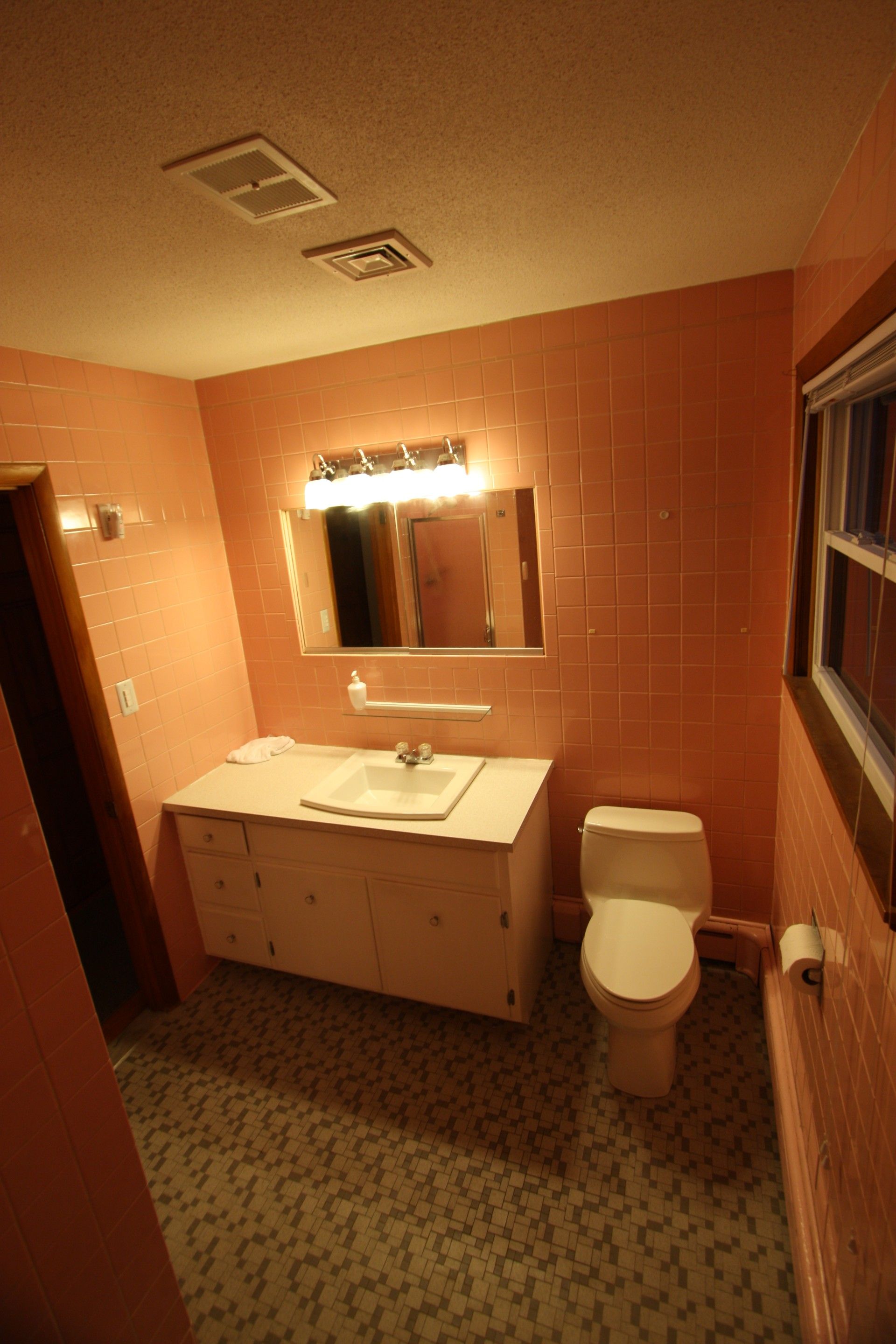 Pink-tiled bathroom with a toilet, sink, mirror, and a small window.