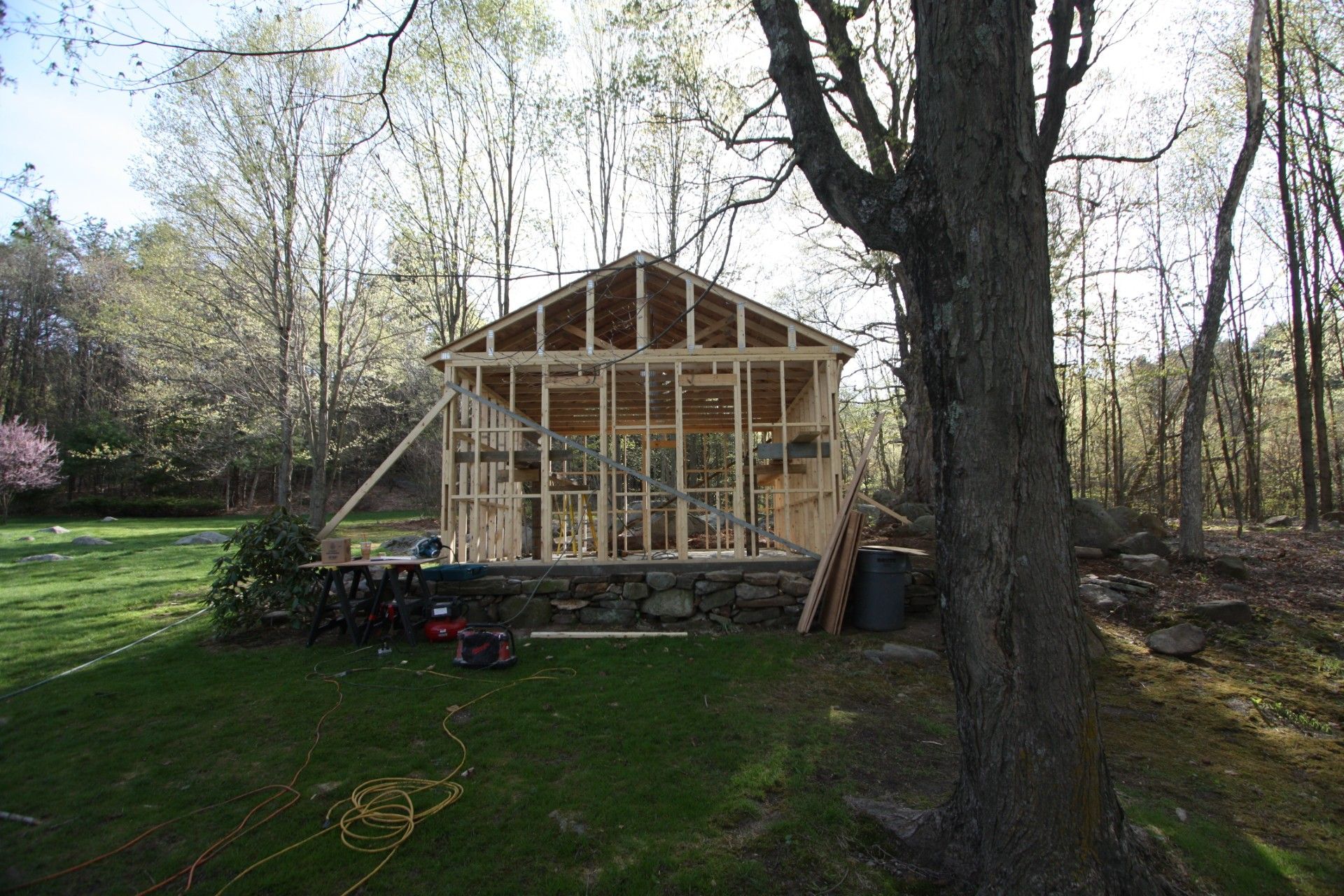 Wood frame structure under construction on a stone base, set in a yard near trees.