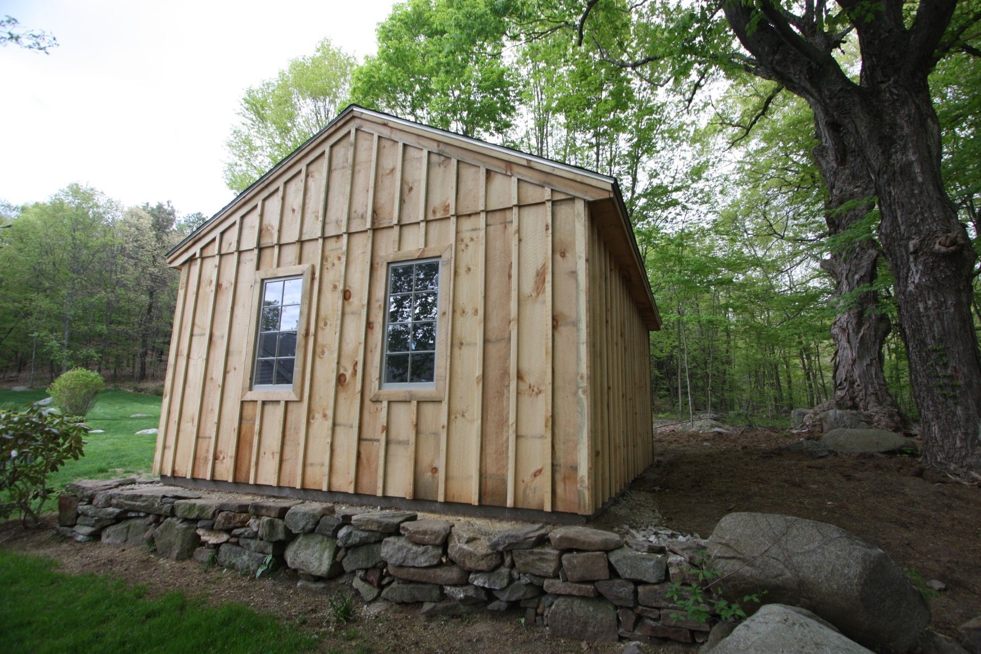 Small, wooden cabin with two windows, set on a stone foundation, surrounded by trees.