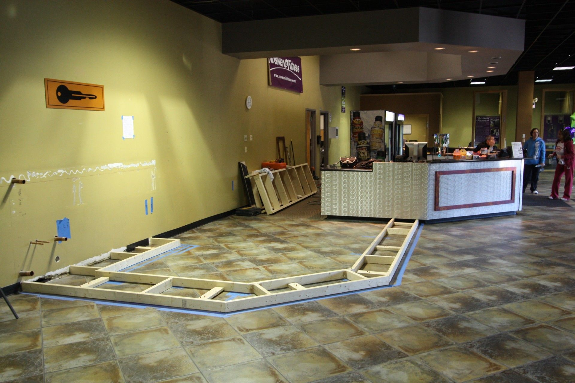 Construction site: wooden frame on floor, beige walls, counter, workers, and building interior.