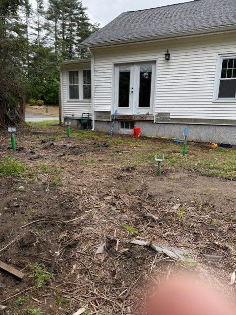 Backyard with bare soil, house with white siding and French doors; green stakes in ground.