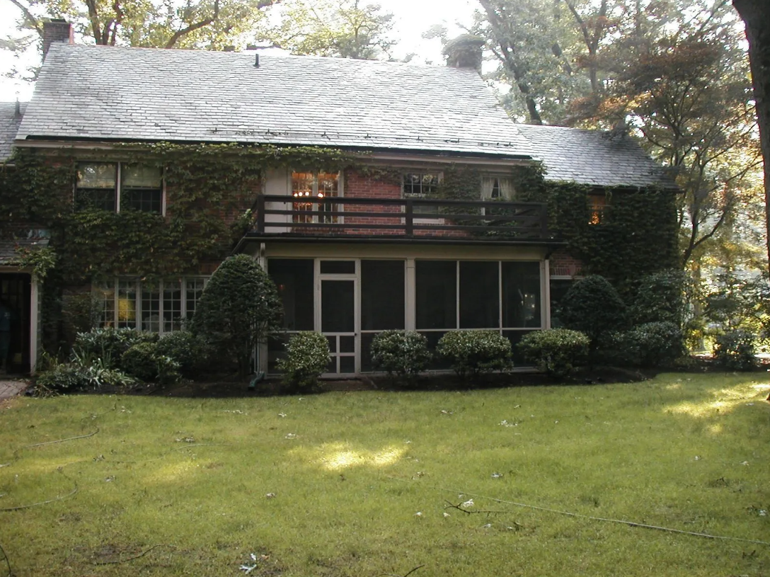 Two-story house with screened porch and balcony, covered in ivy, set in a yard with trees.
