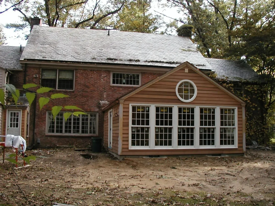 Red brick house with wooden sunroom addition, surrounded by trees and a dirt yard.