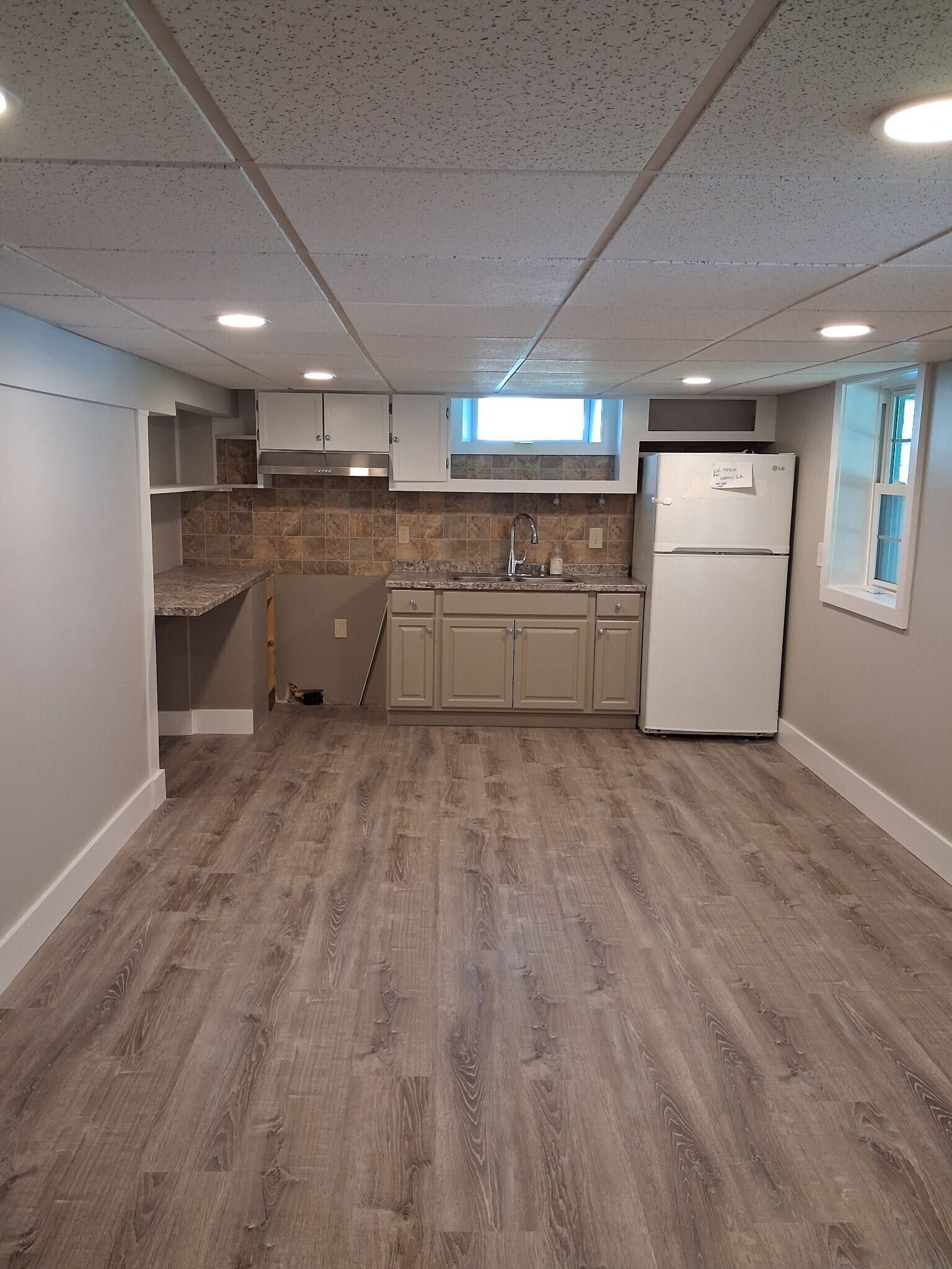 Empty basement kitchenette with beige cabinets, white fridge, wood-look flooring, and a small window.