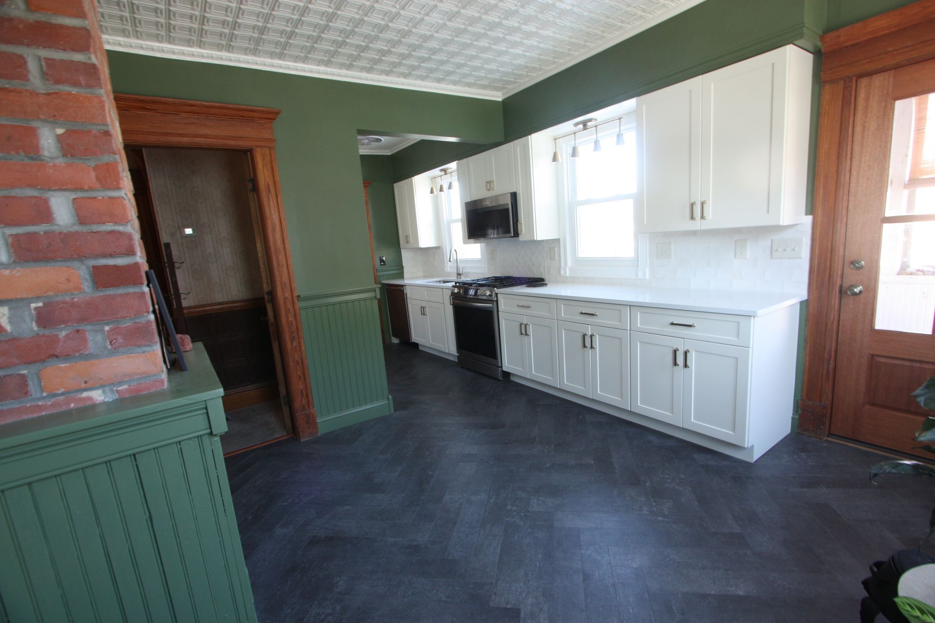 Empty kitchen with white cabinets, black tile floor, green walls, and a brick fireplace edge.