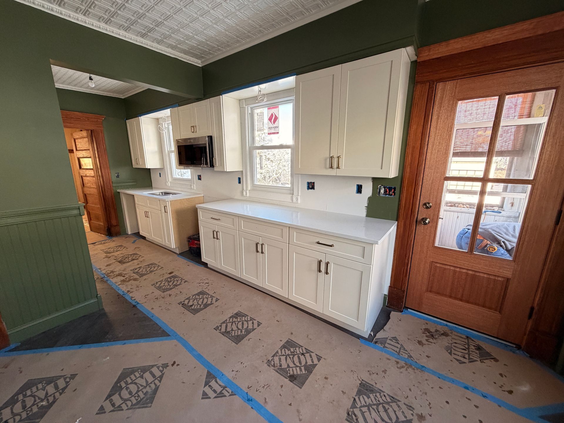 Kitchen with white cabinets, green walls, wood door, and taped-off floor during renovation.