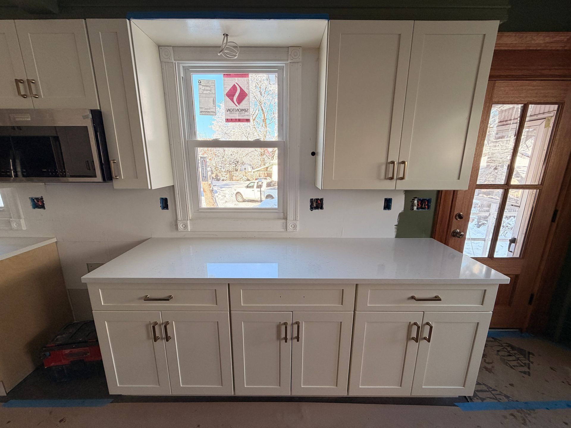 White kitchen cabinets and countertop beneath a window, with a microwave on the left and wood door on the right.
