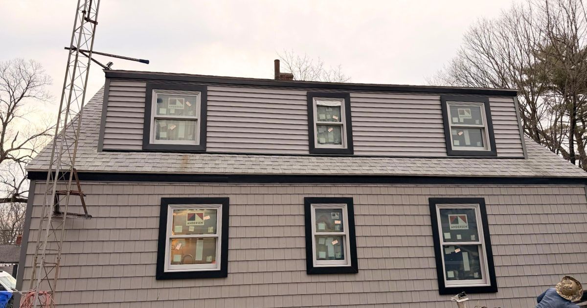 Gray house exterior with dark-trimmed windows and a sloped roof under a cloudy sky