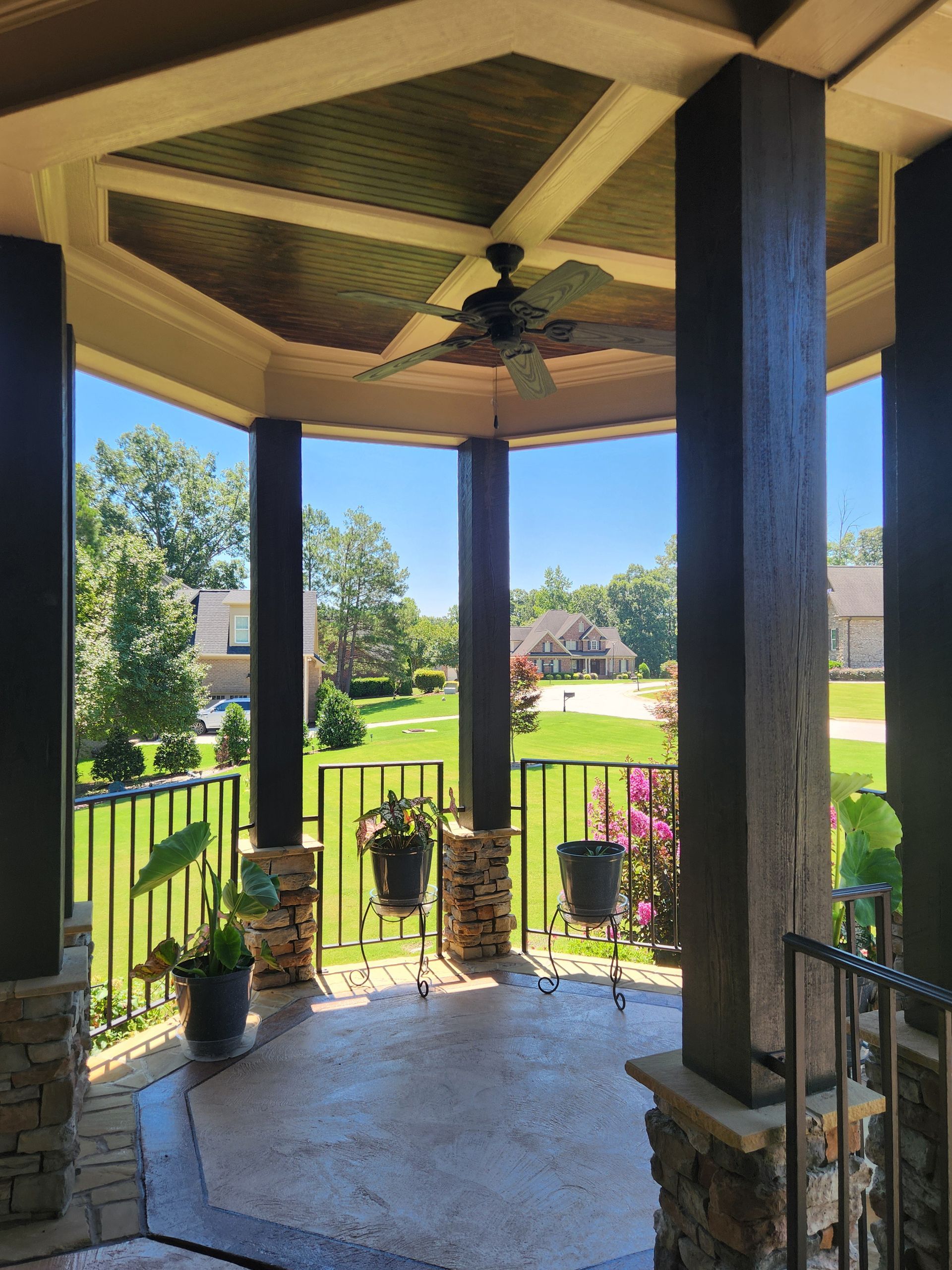 Covered porch with ceiling fan, dark columns, and potted plants overlooking a sunny green lawn and garden