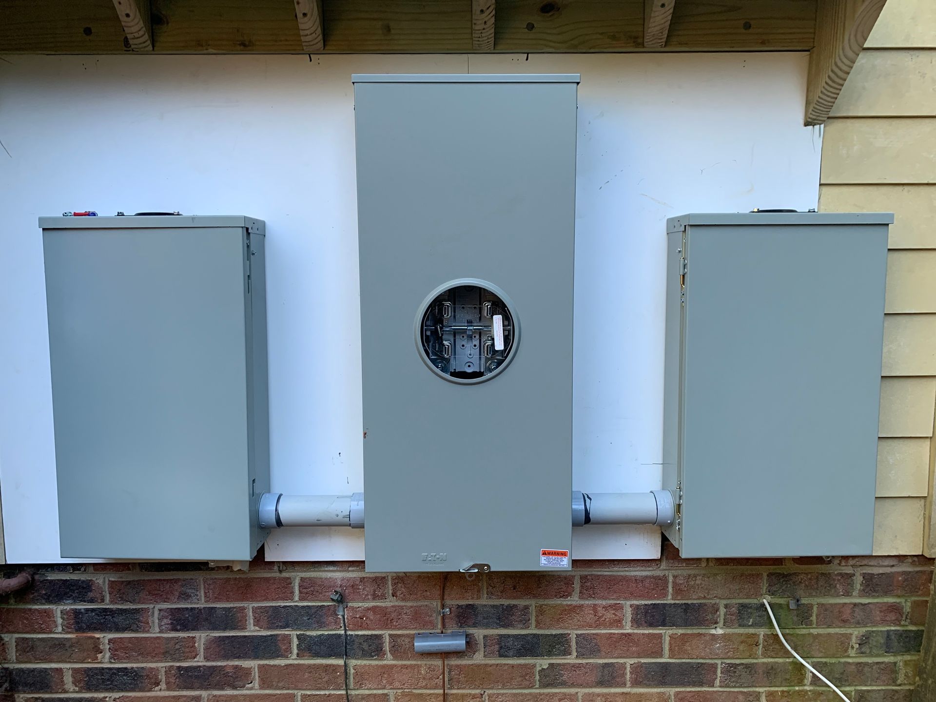 Three gray electrical meter boxes mounted on a brick wall under a roof eave
