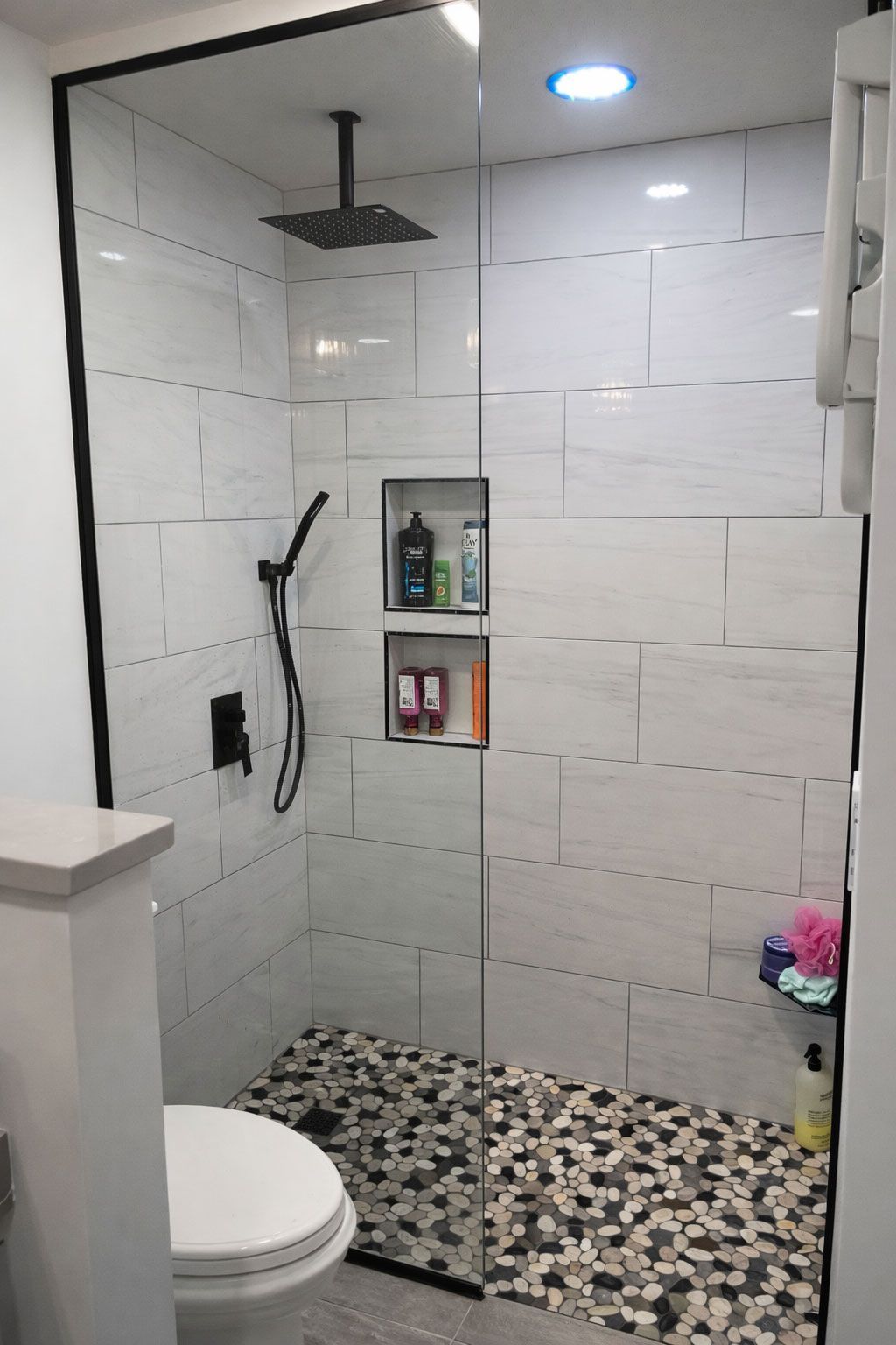 Small white-tiled bathroom shower with black fixtures, pebble floor, and recessed shelf.