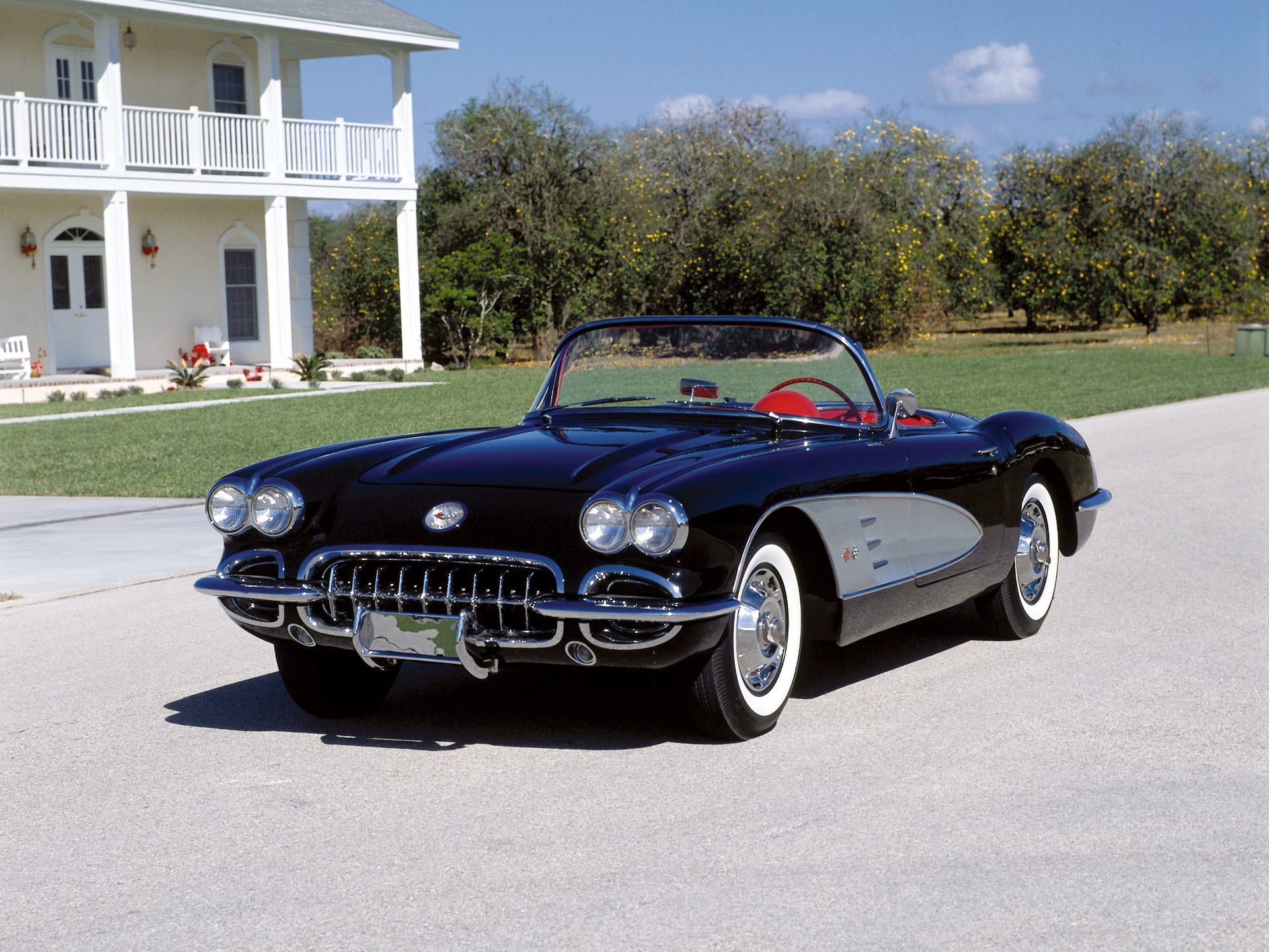 Black vintage convertible car parked on a paved road, white-walled tires, red interior, house in background.