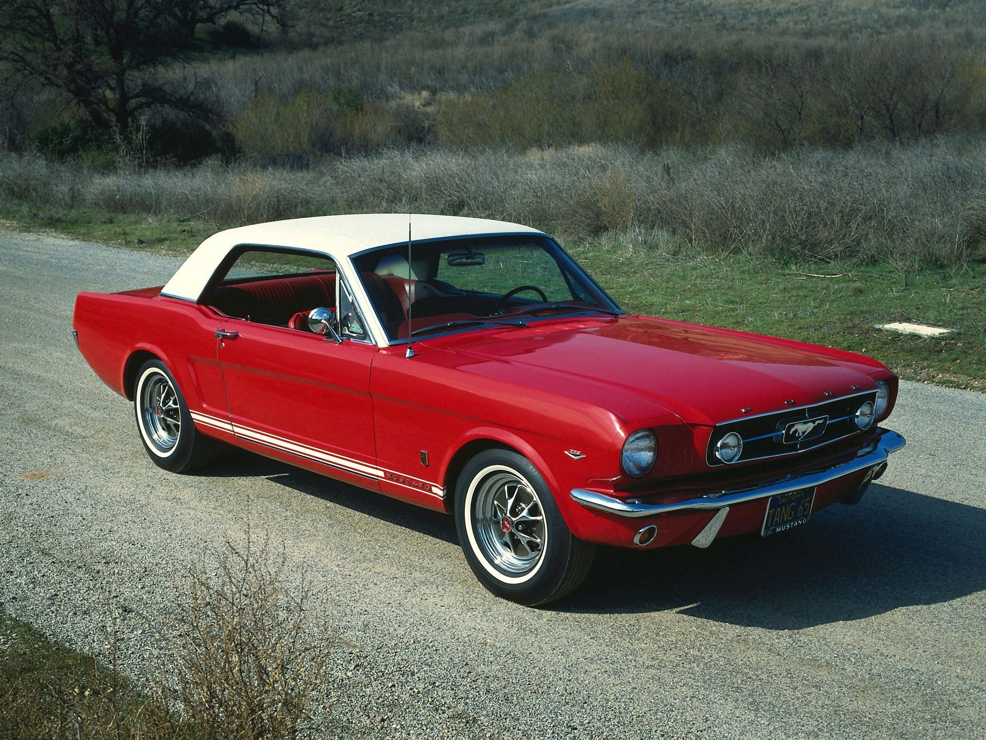 Red 1965 Ford Mustang coupe with a white roof on a gravel road, parked on a roadside with vegetation.