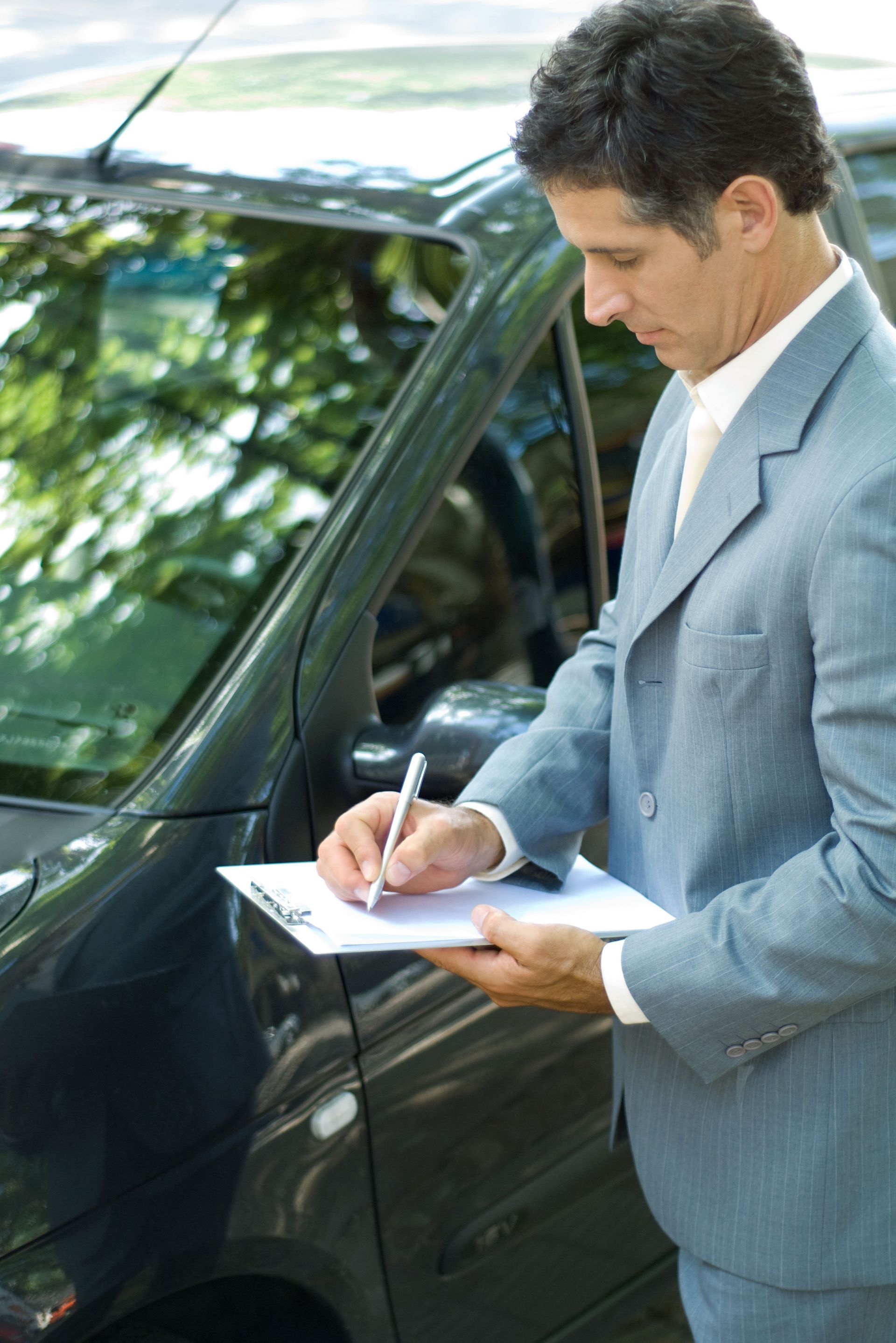 Man in suit writing on clipboard next to a dark car.