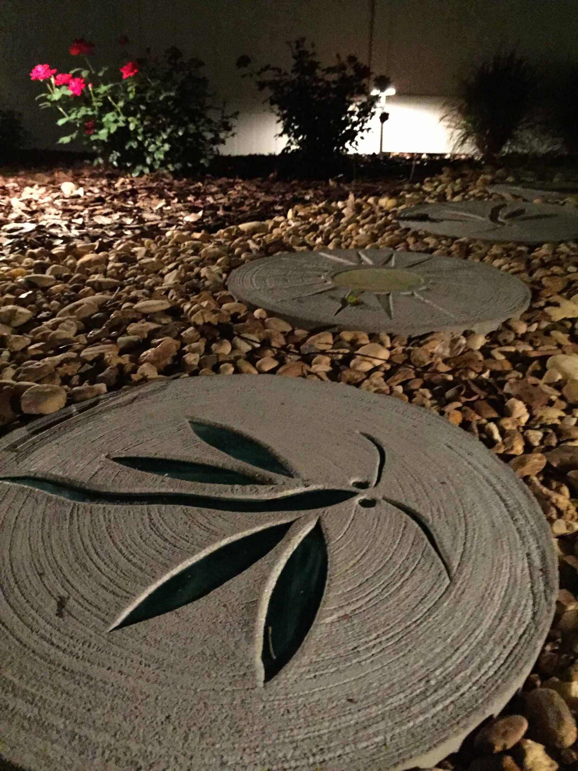 Stone pathway with leaf designs lit up at night, surrounded by rocks and bushes.