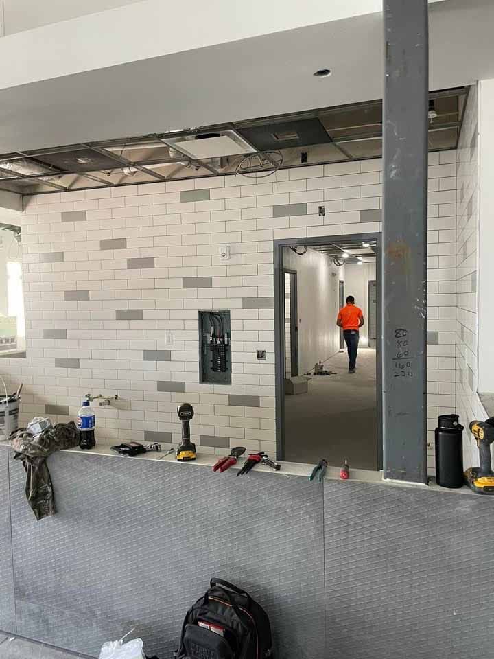 Construction site interior: man walks down hallway. Walls have white and grey tile. Tools and equipment on countertop.