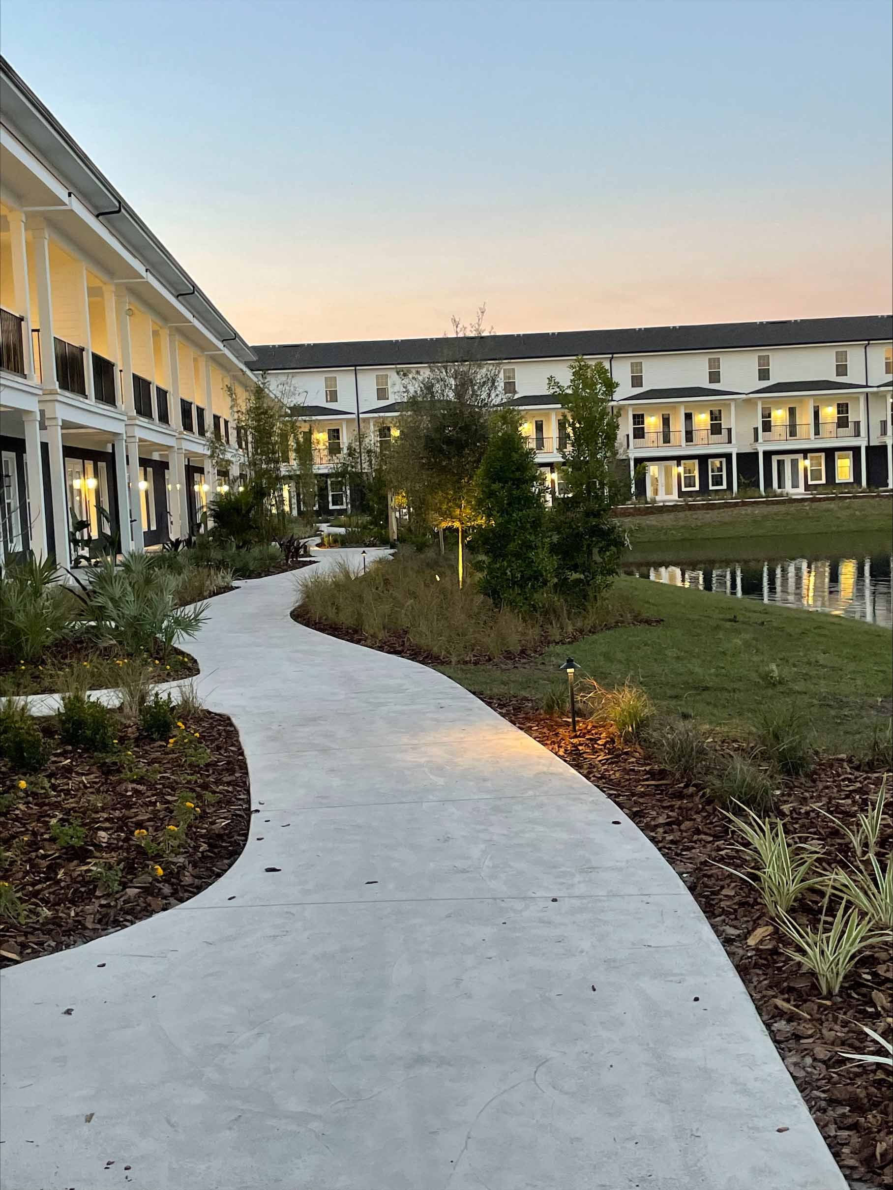 Paved pathway through landscaped area to buildings near a lake. Dusk lighting.