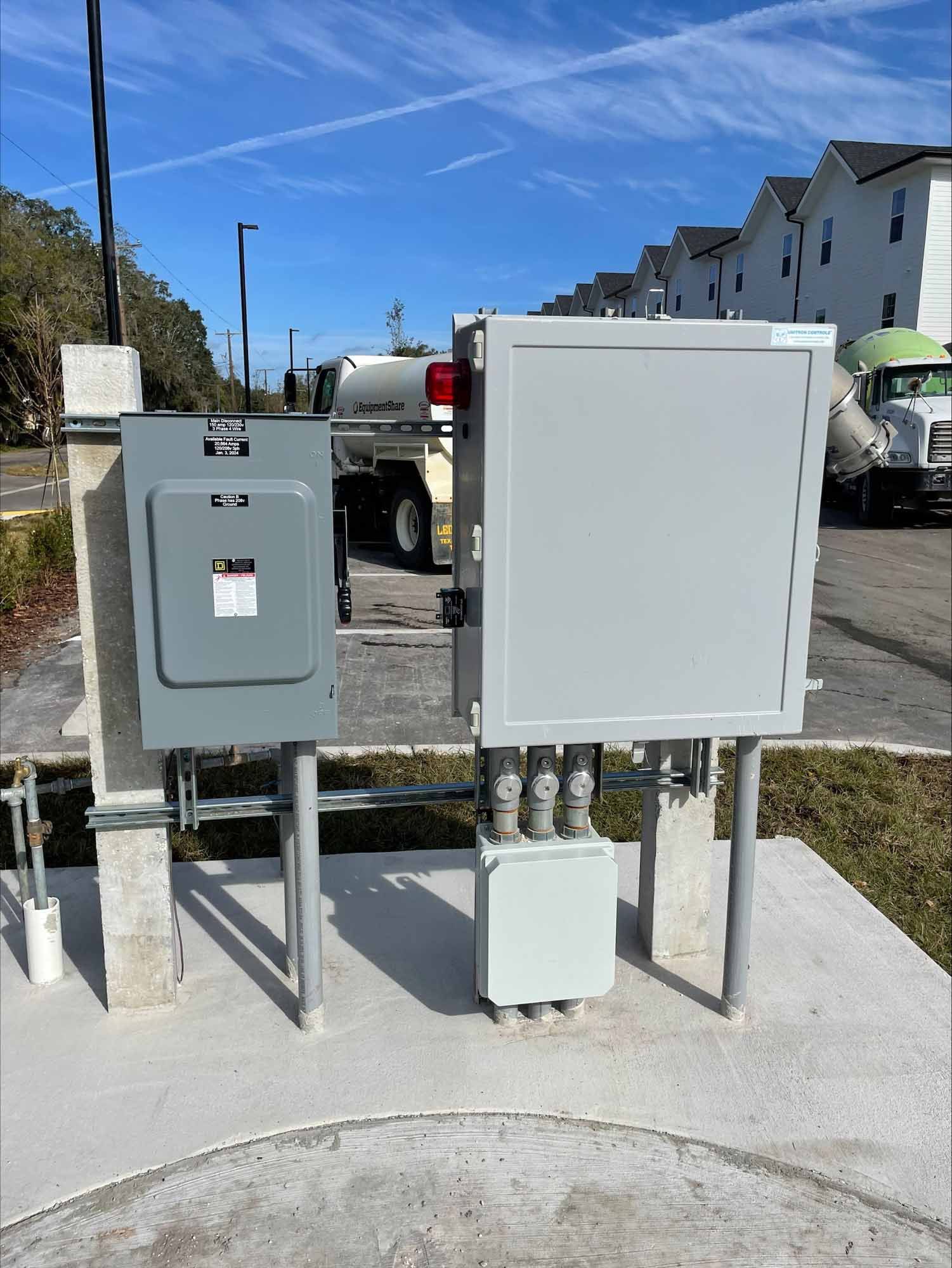 Gray electrical boxes on concrete pad, utility trucks in background, blue sky.