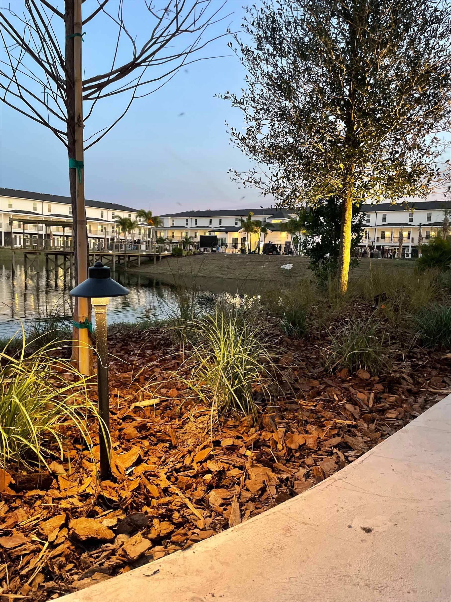 Lit landscape light near a pond, with townhouses in the background under a dusky sky.