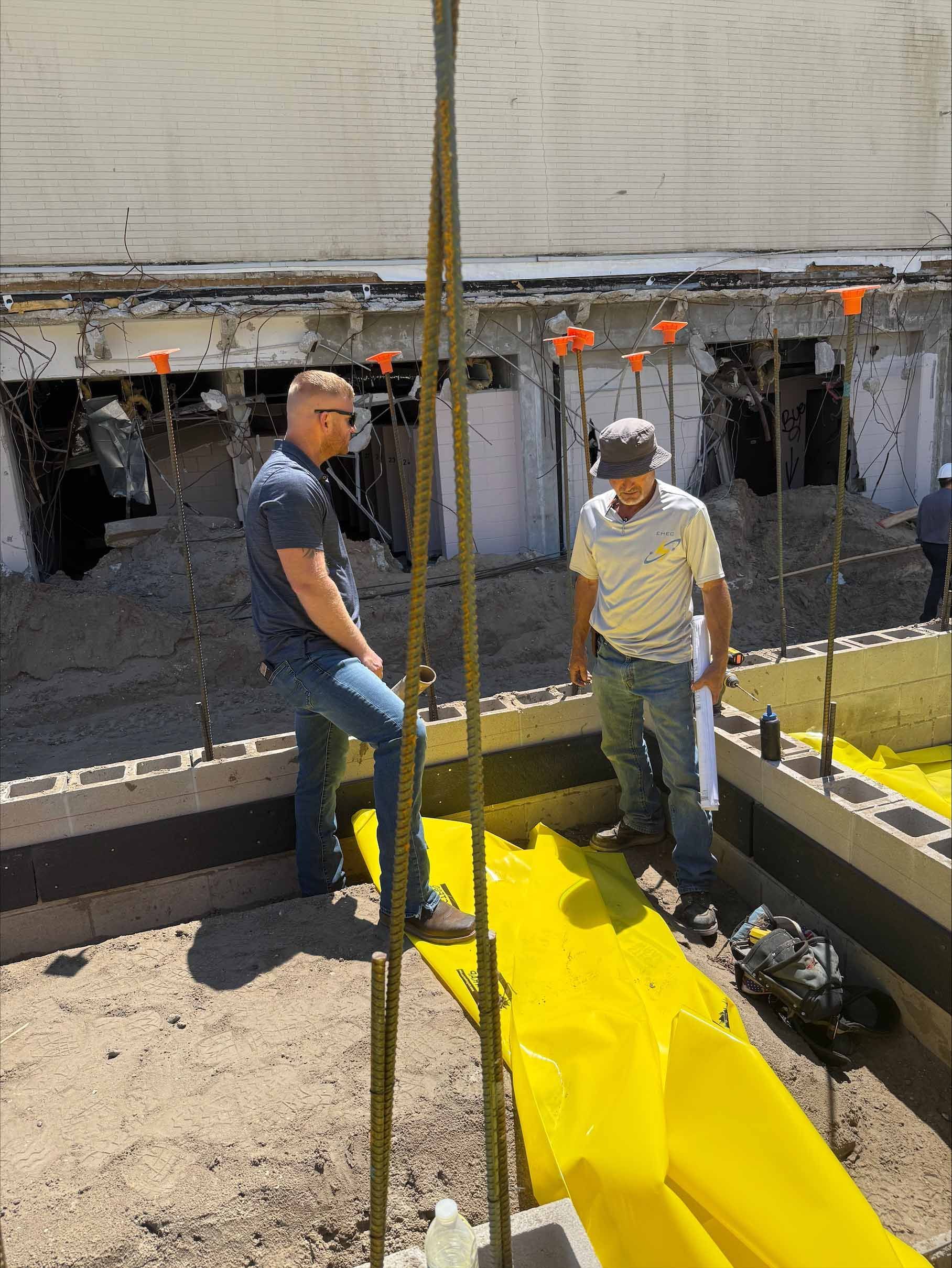 Two men examining a construction site with rebar, blocks, and yellow tarps. Outdoors, sunny.
