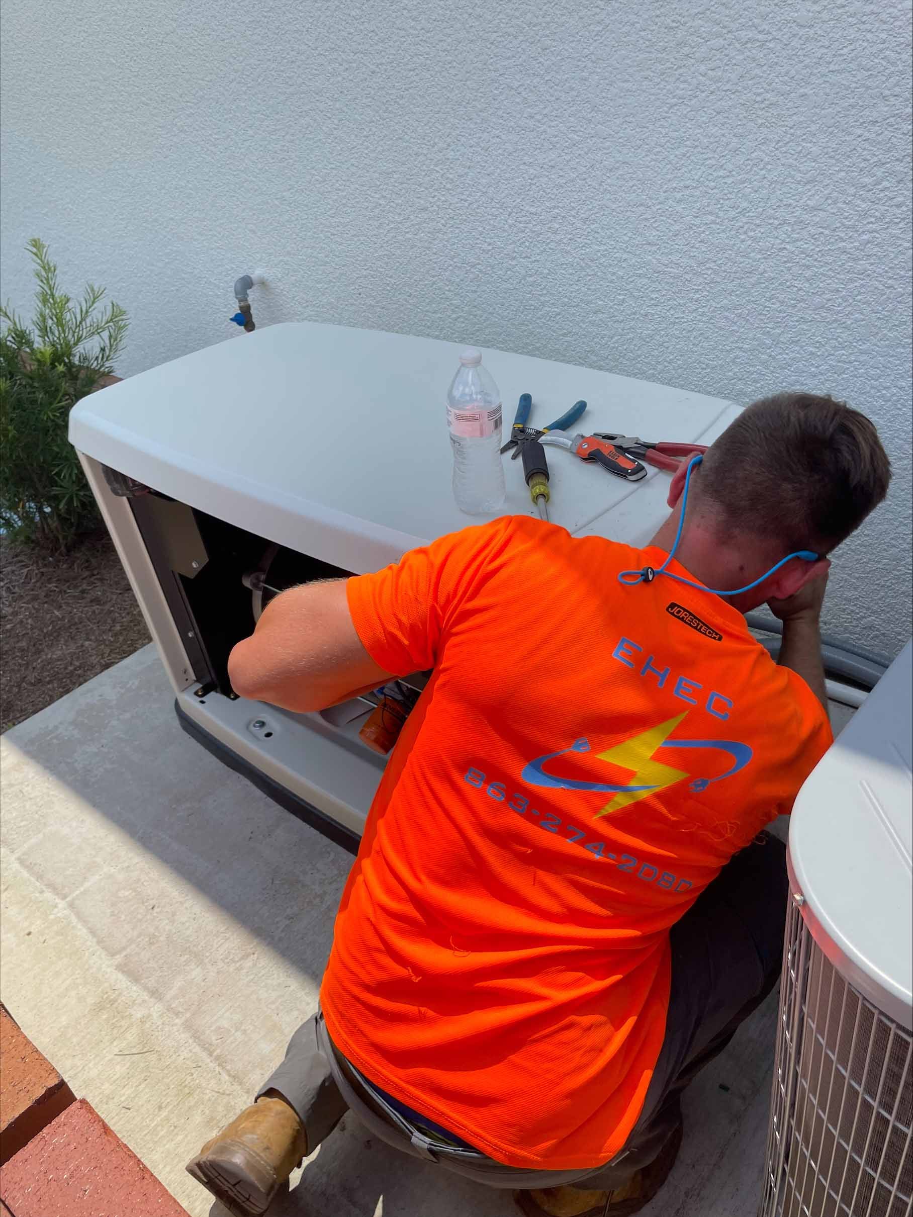 Man in orange shirt repairs a generator outdoors, kneeling on concrete. Tools and water bottle visible.
