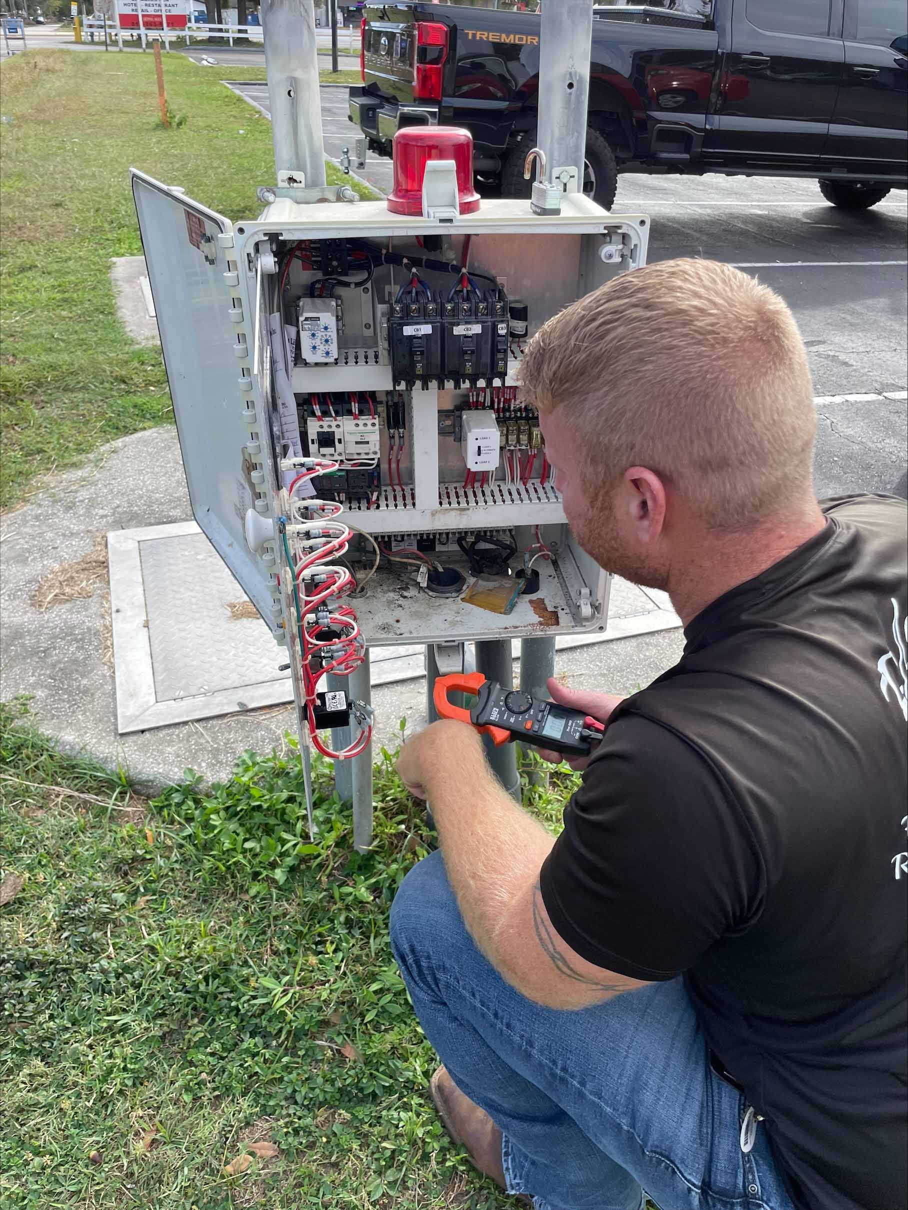 Man working on electrical equipment outdoors, kneeling, using a tool.