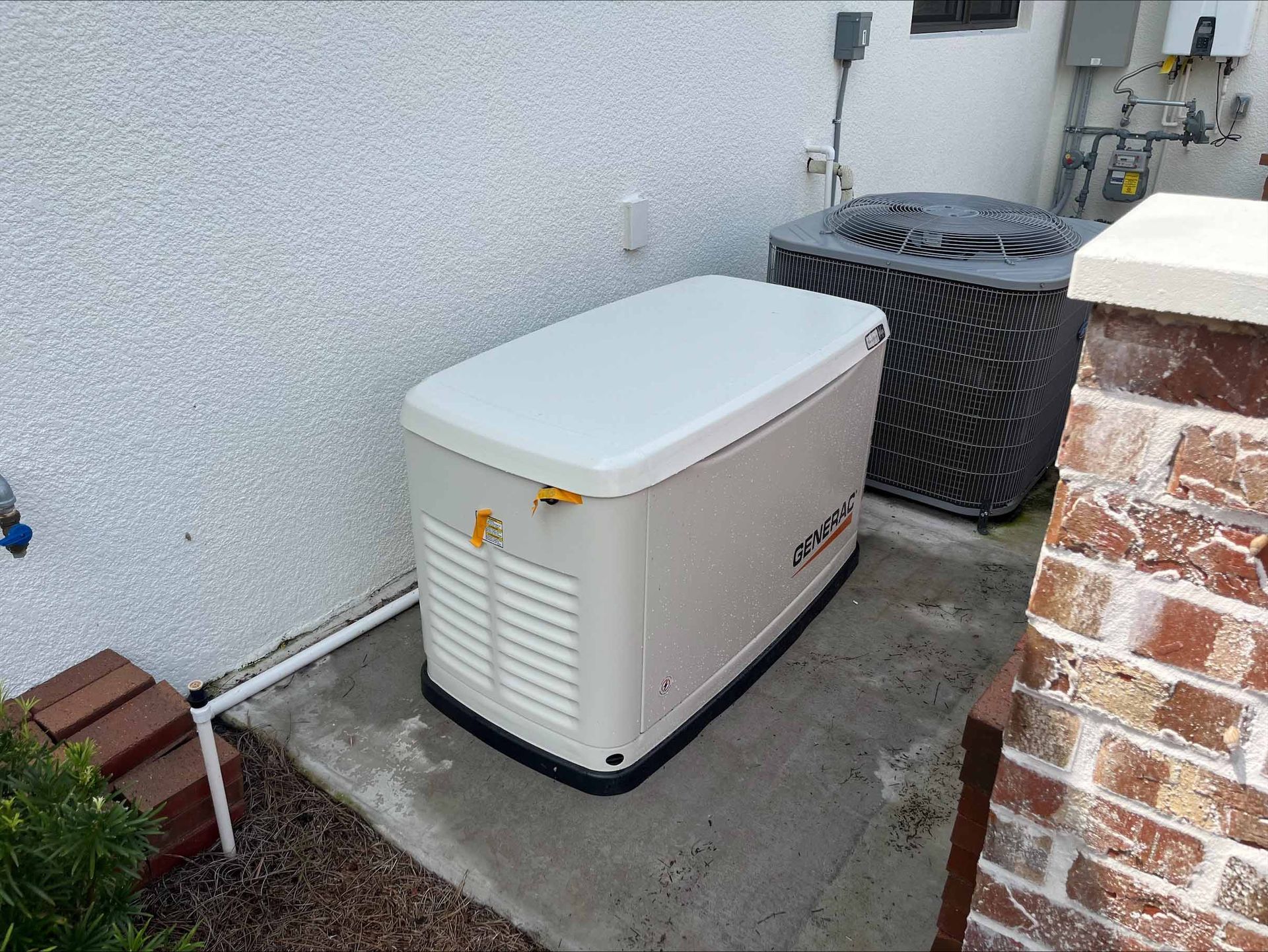 A beige home generator and black AC unit sit outside on a concrete pad next to a brick wall.