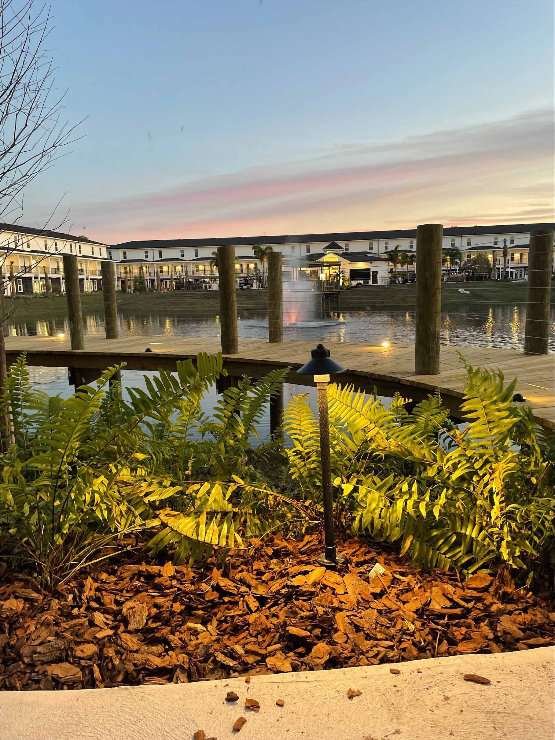 Wooden dock with landscape lighting over a pond, with a fountain and buildings in the background. Evening sky.