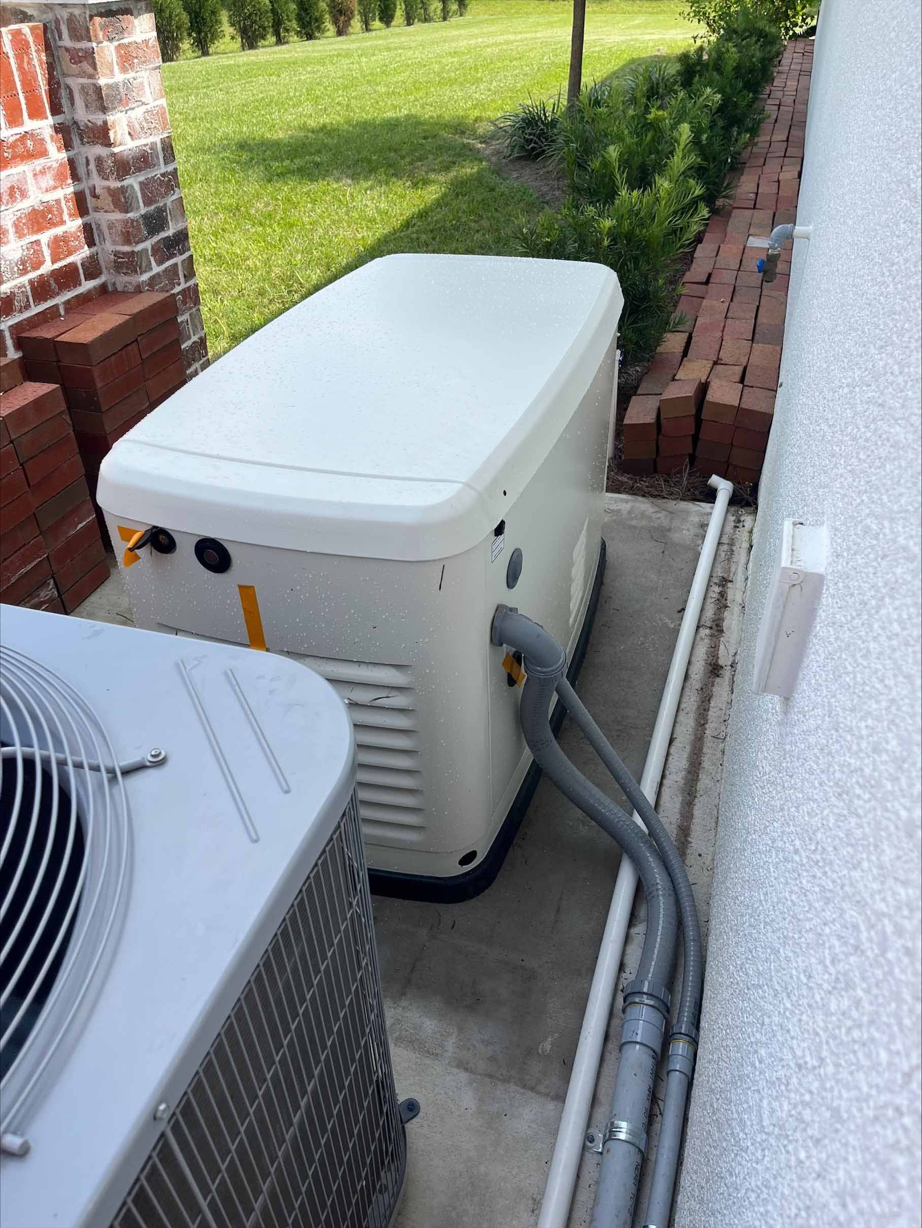 A generator sits next to an air conditioning unit near a brick wall and hedge.