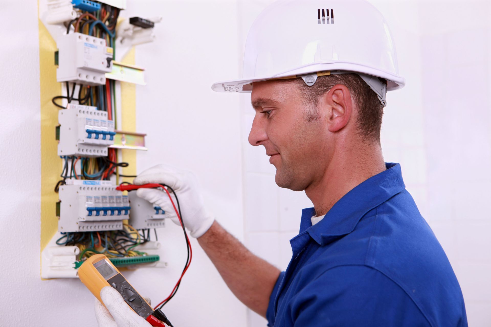 Electrician in blue shirt and white hard hat uses a multimeter on a circuit panel.