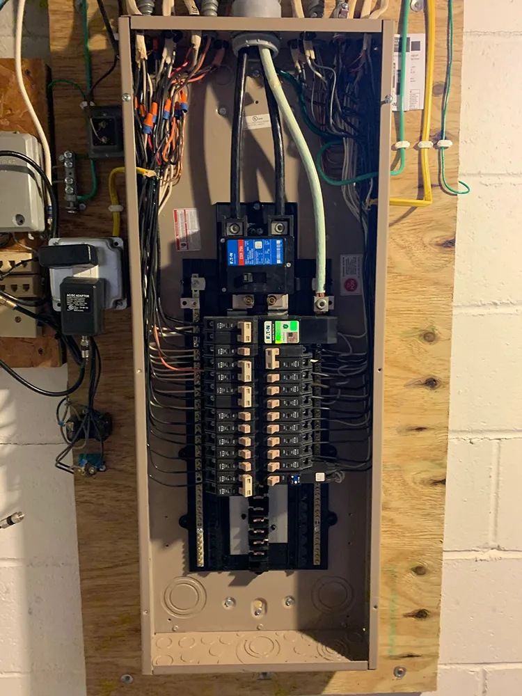 Electrical panel mounted on a wooden board against a wall, with wires and circuit breakers visible.
