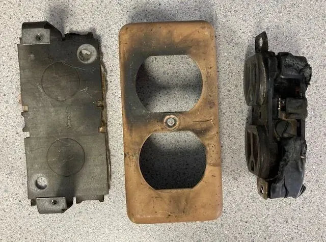 Burnt electrical outlet parts: metal bracket, faceplate, and wiring component, showing damage.
