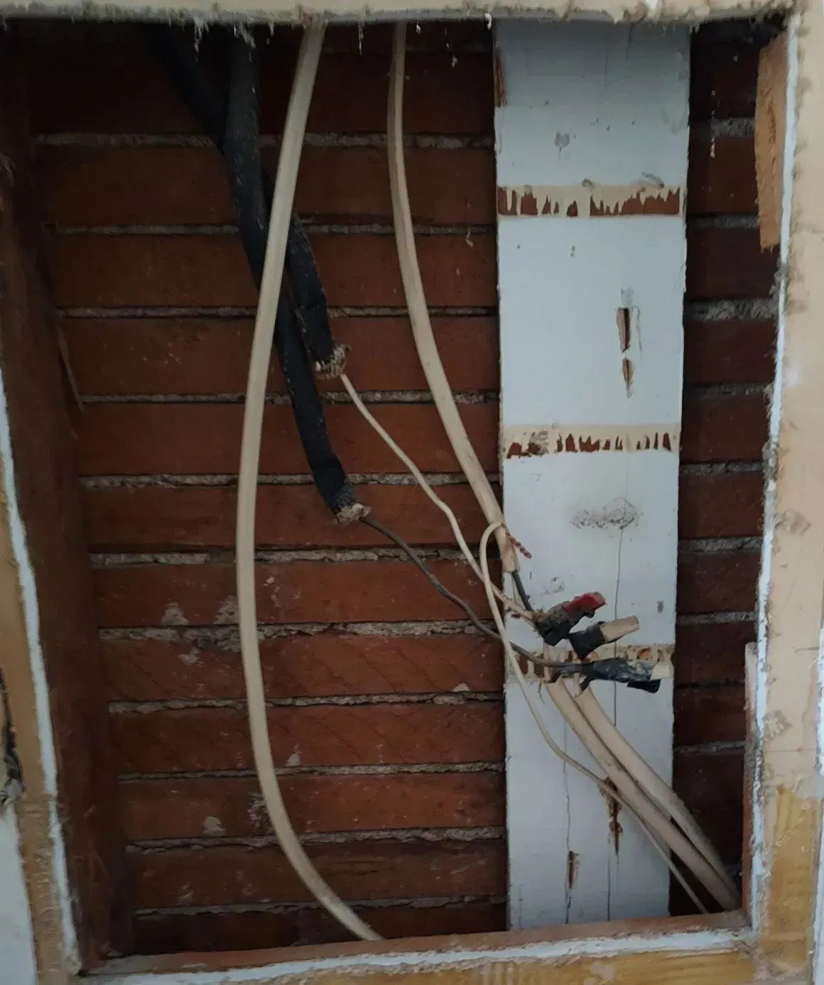 Exposed electrical wiring and pipes in a wall cavity, with a wooden backing and brick.