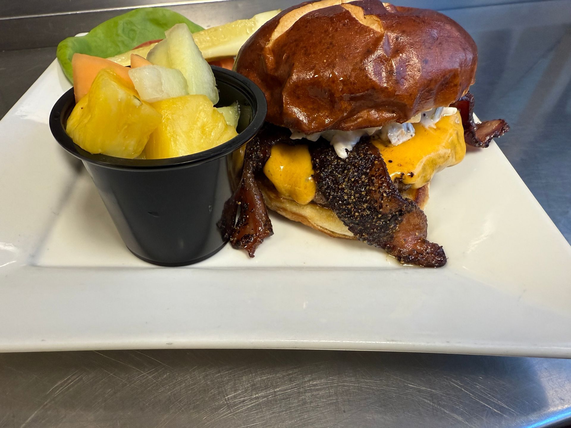 Burger with bacon, cheese, and pretzel bun, served with fruit salad.
