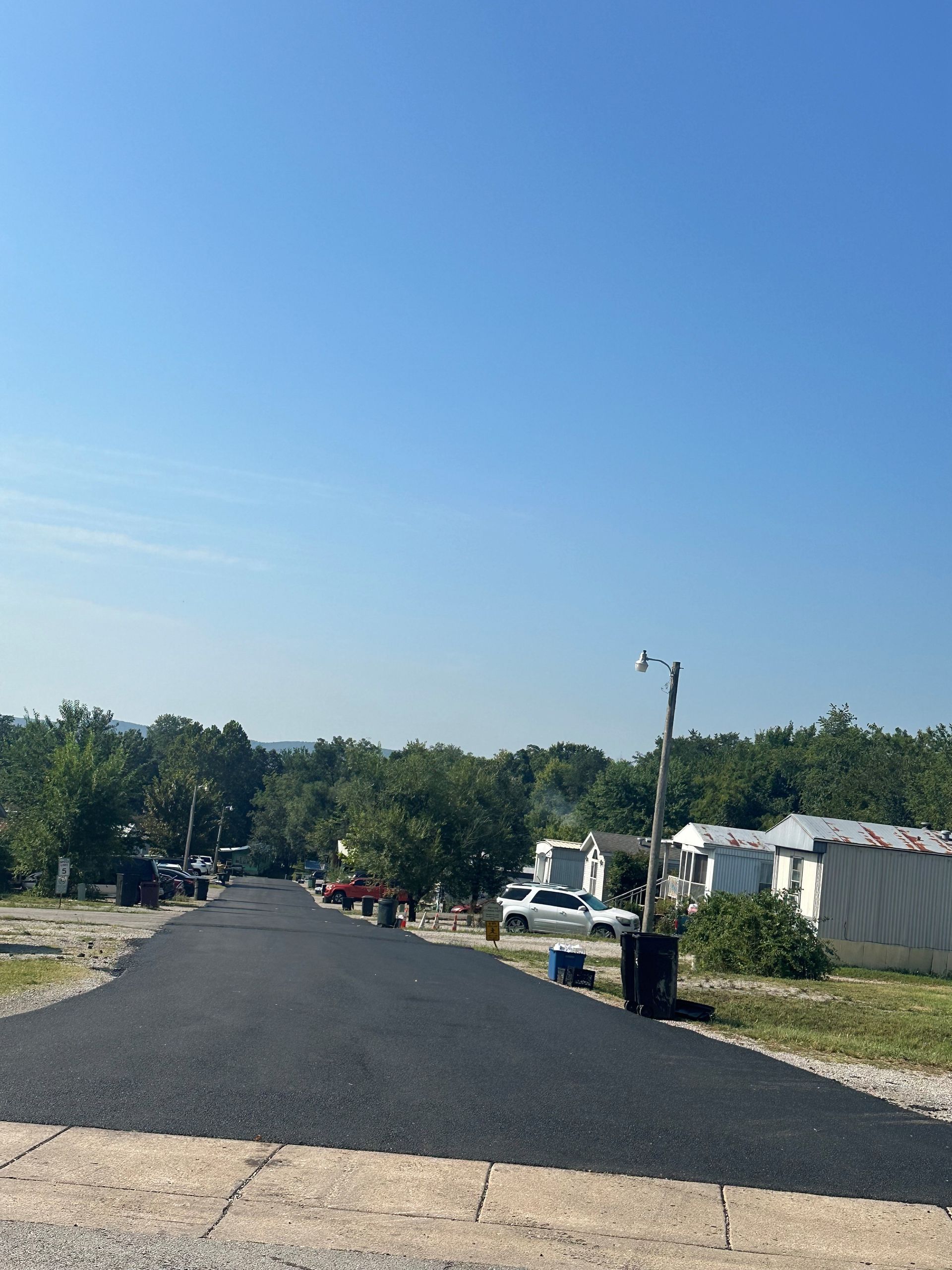 Paved road in a mobile home park on a sunny day, with trees, trailers, and a few people in the distance