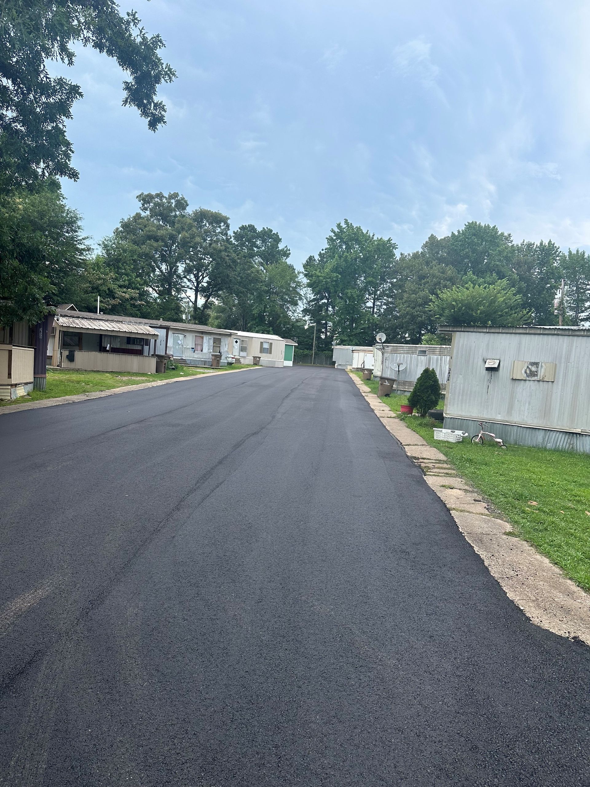 Freshly paved road in a mobile home park lined with white trailers and trees under a cloudy sky