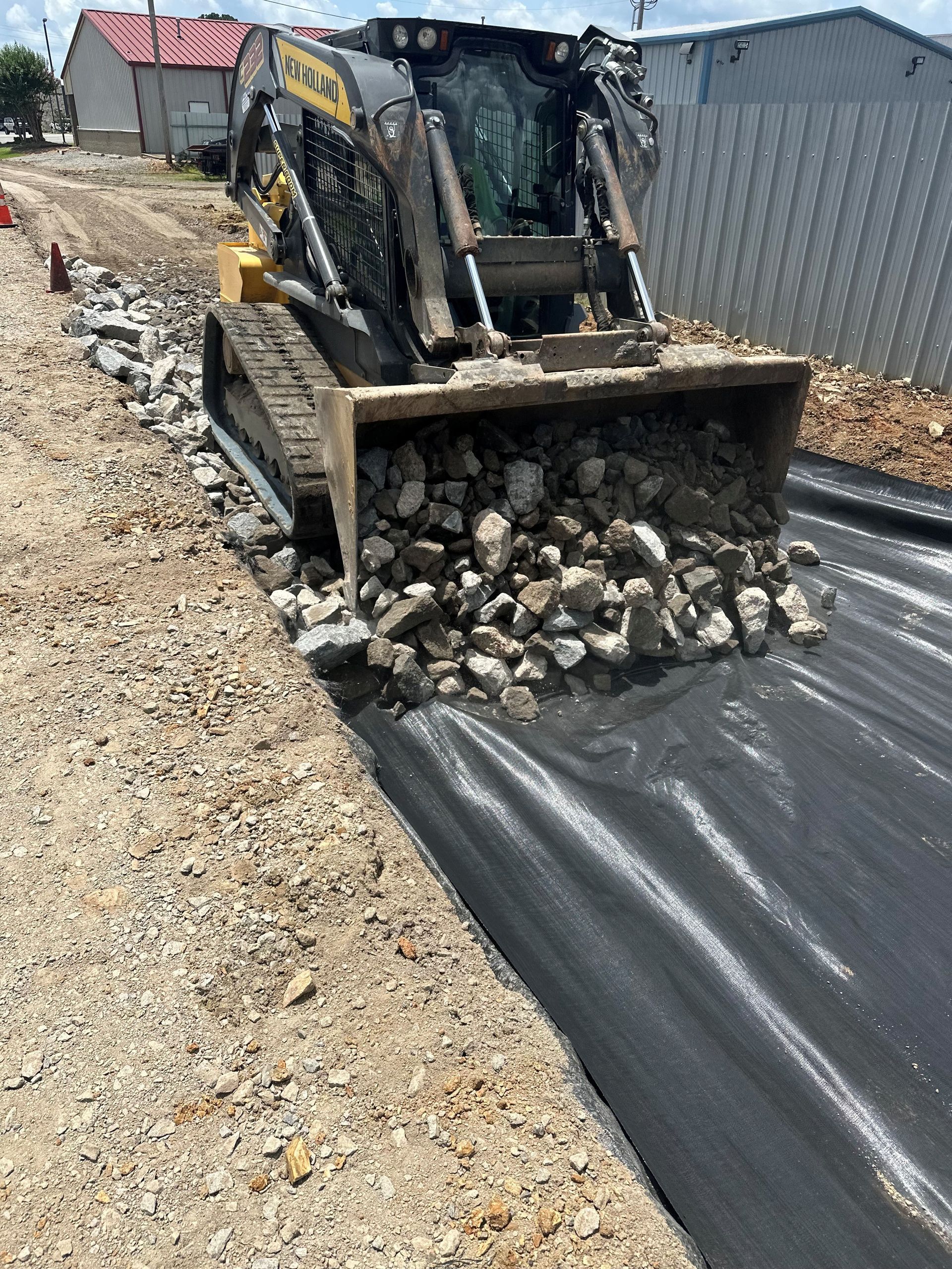 Backhoe dumping rocks onto black landscaping fabric beside a gravel path