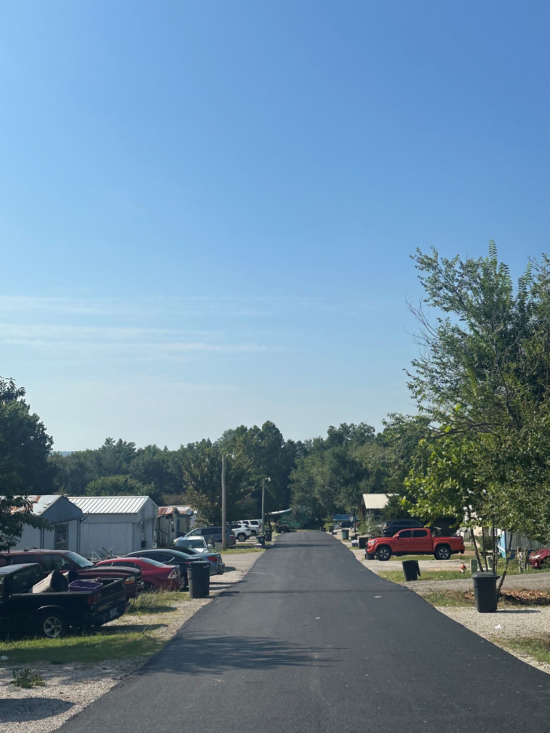 Quiet mobile home park street under a clear blue sky, with parked cars and trees lining both sides.
