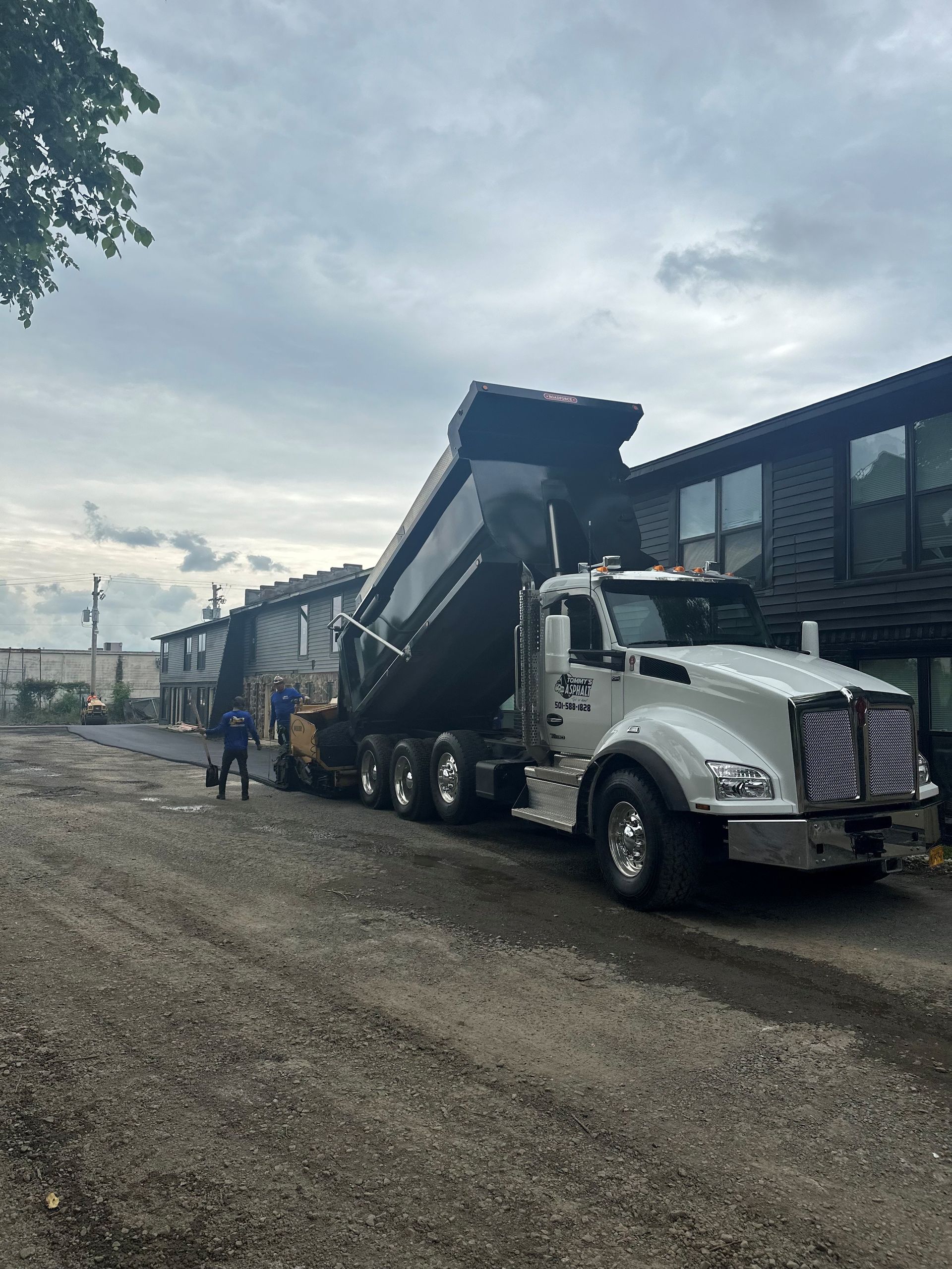 White dump truck with raised bed parked on a gravel lot beside a dark building under cloudy skies