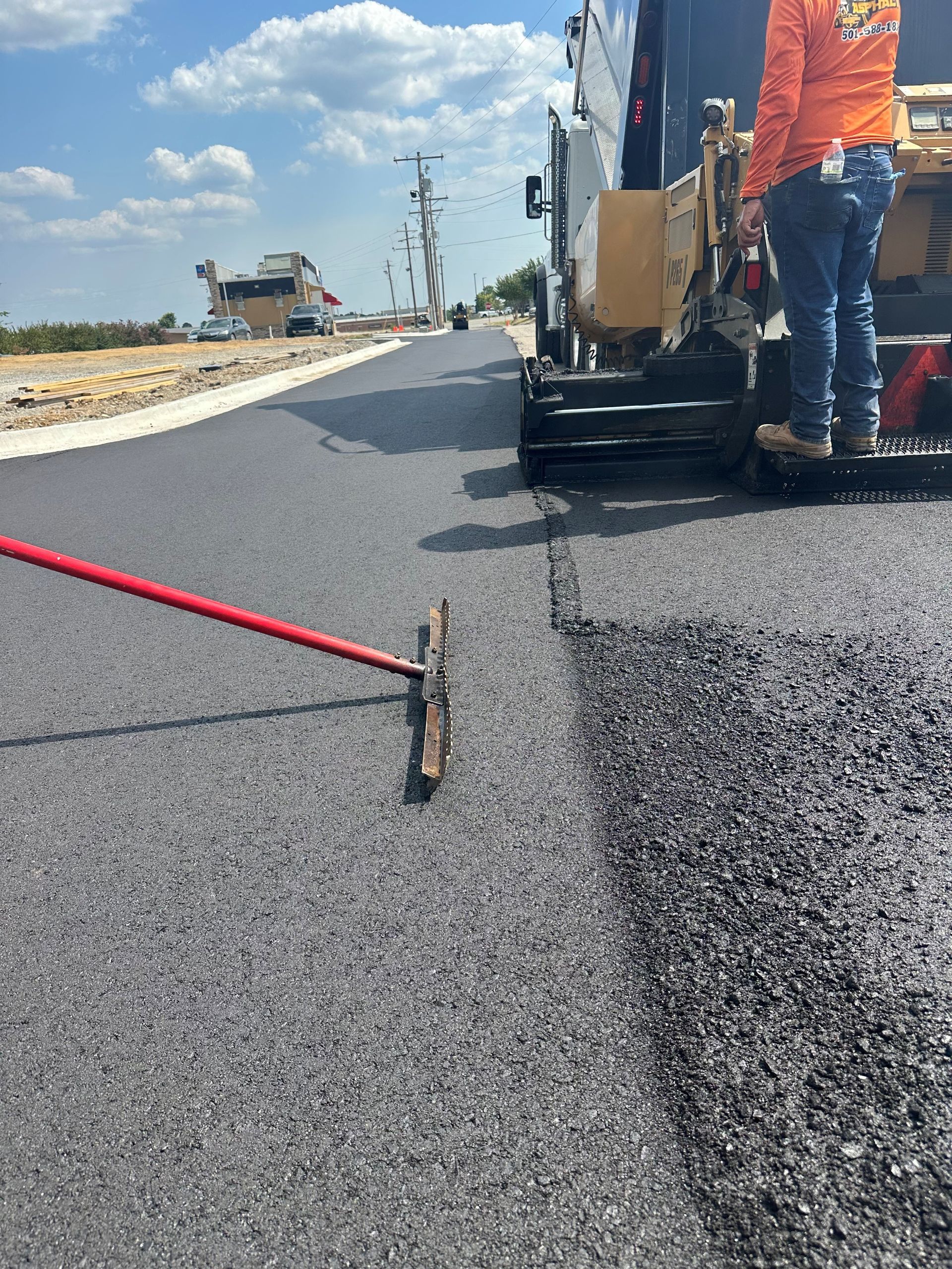 Road construction worker smoothing fresh asphalt beside a paving machine on a sunny street