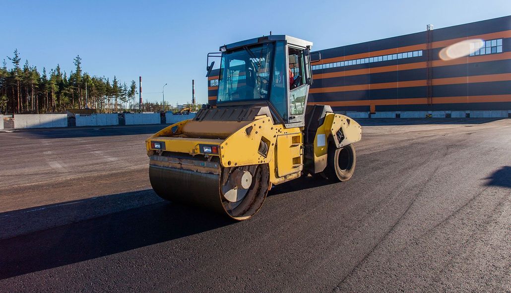 Yellow road roller on a paved lot near a large industrial building under clear blue sky