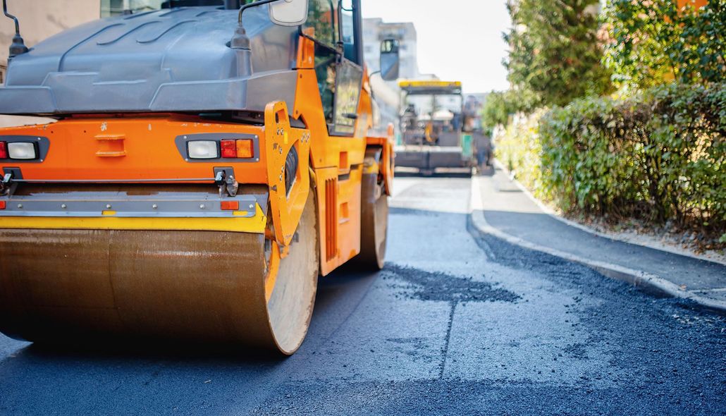 Orange road roller paving a fresh asphalt street beside a hedge